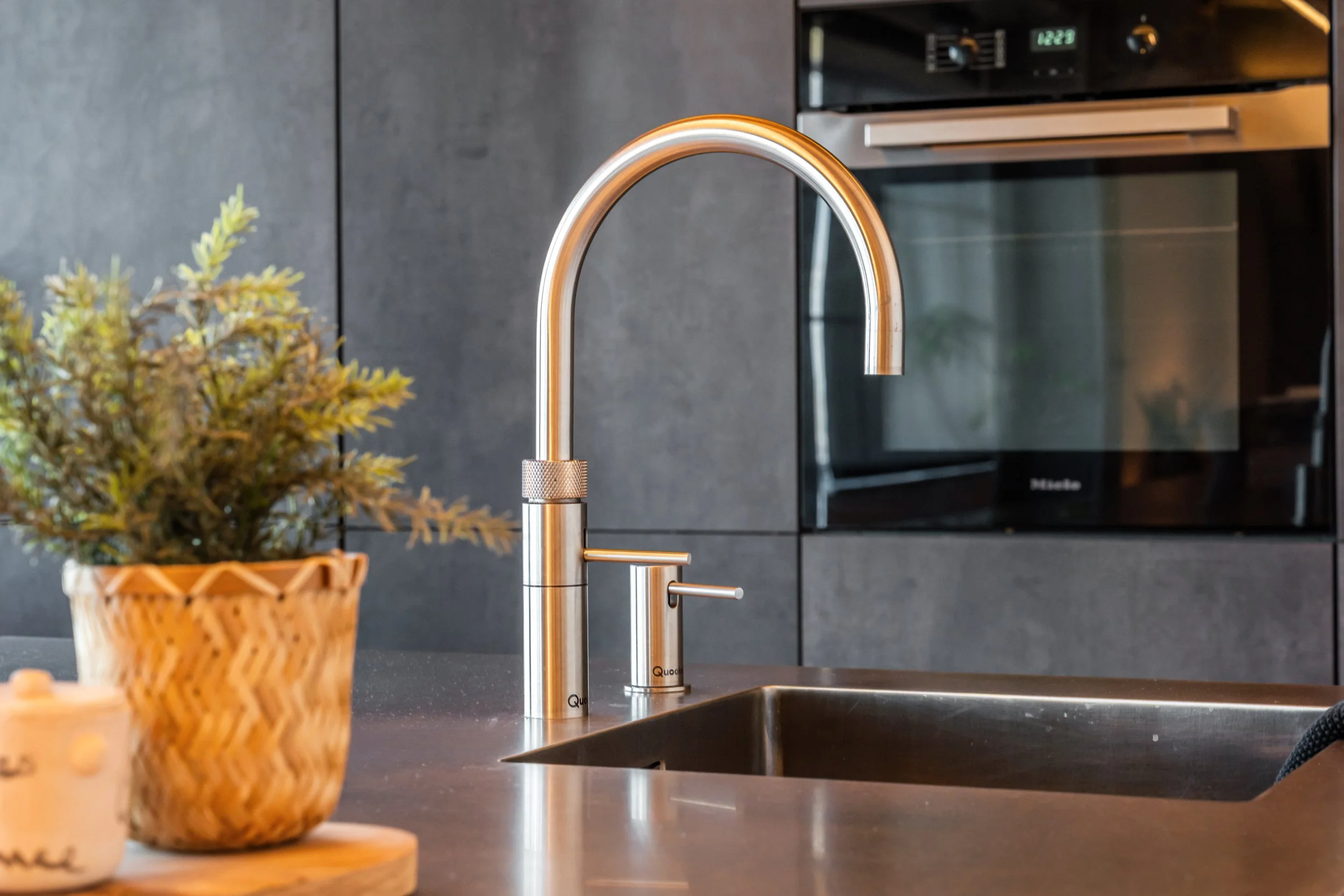 Een moderne keuken met een strak aanrecht, een gloeilamp-achtige keukenkraan, en een plantenpot op het aanrecht.