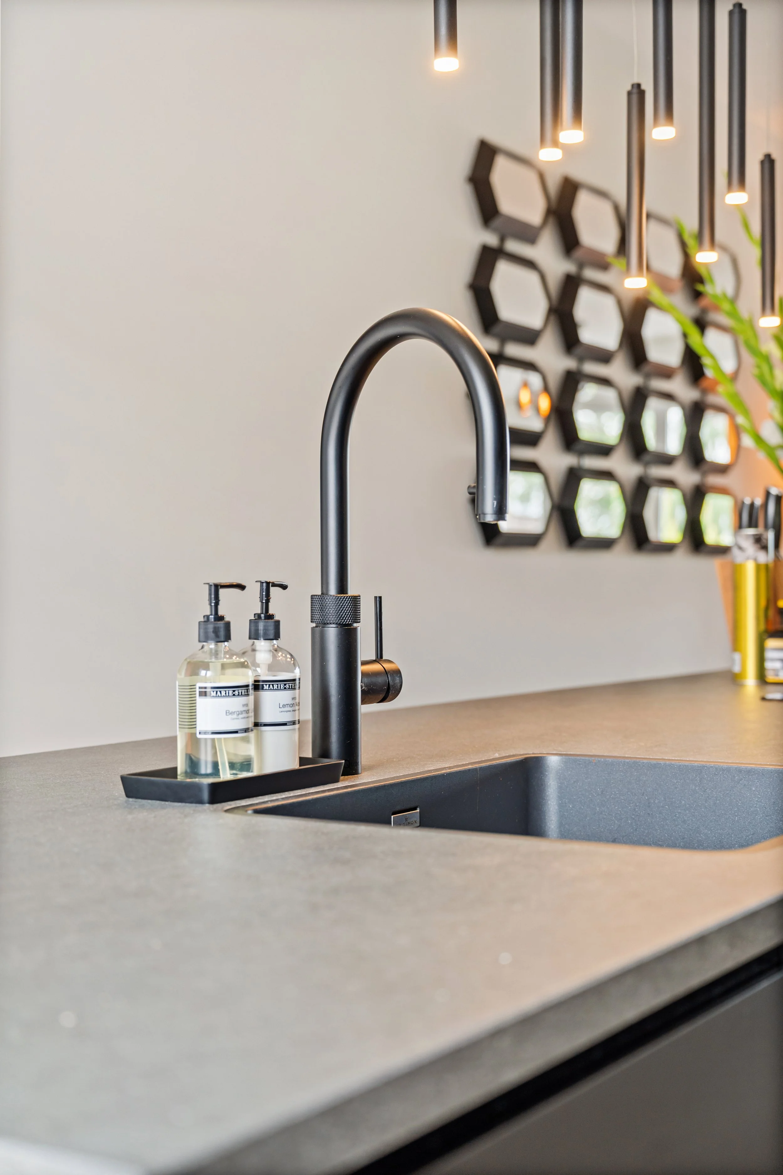 Een modern keukenaanrecht met een zwarte kraan, twee zeepdispensers, en een decoratief muurrek met hexagonale spiegels. Er is een plant in een pot aan de rechterkant.