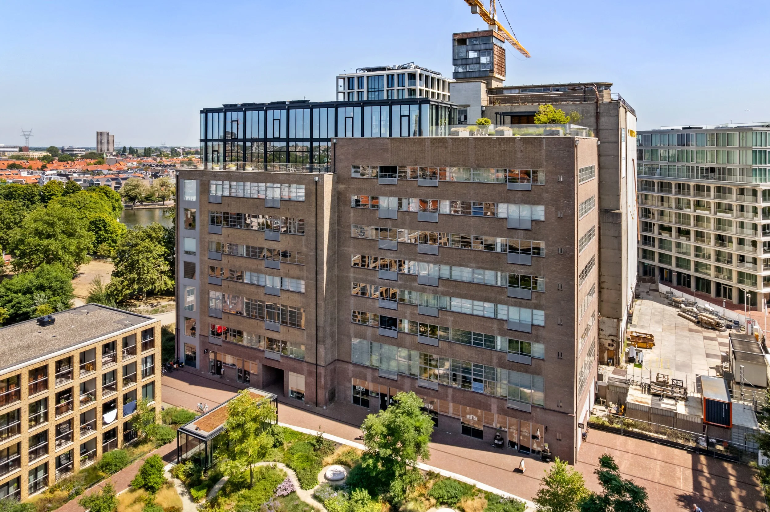 Hoog gebouw onder renovatie met een bouwkraan erboven. Op de begane grond bevinden zich winkels en een aangelegd tuin met bomen en bloemen.