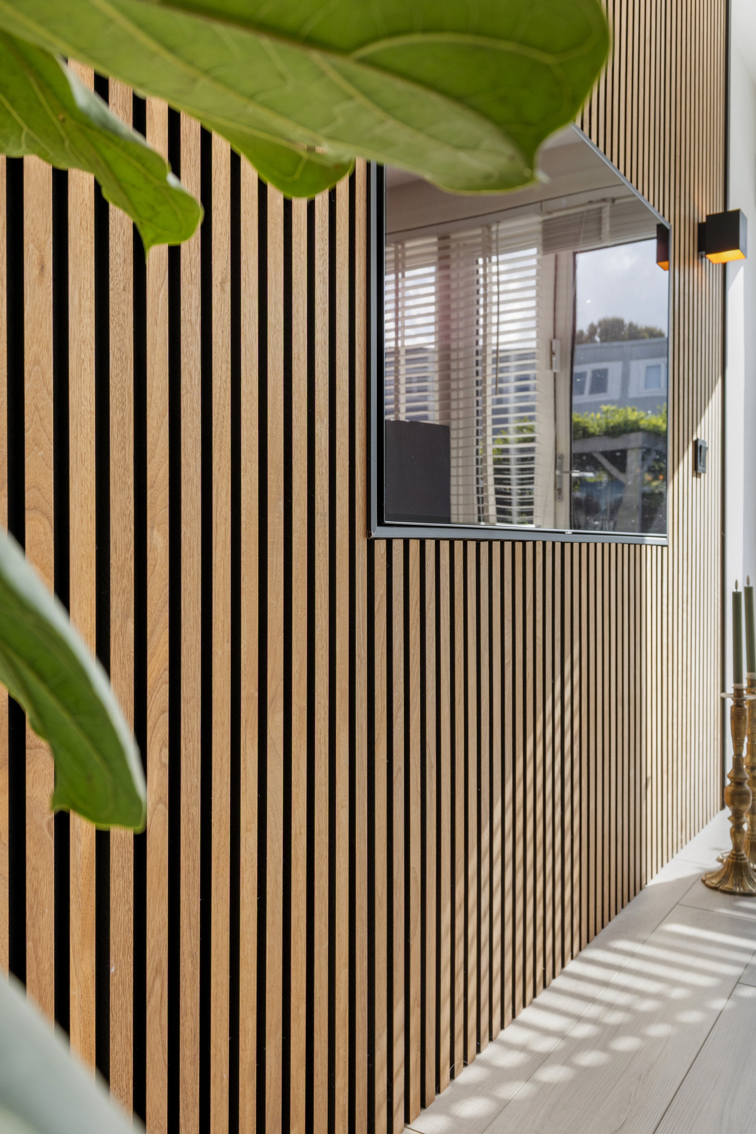 Interieur met een houten wand met verticale latten, een grote spiegel, een plantenblad in de voorgrond en een houten vloer.