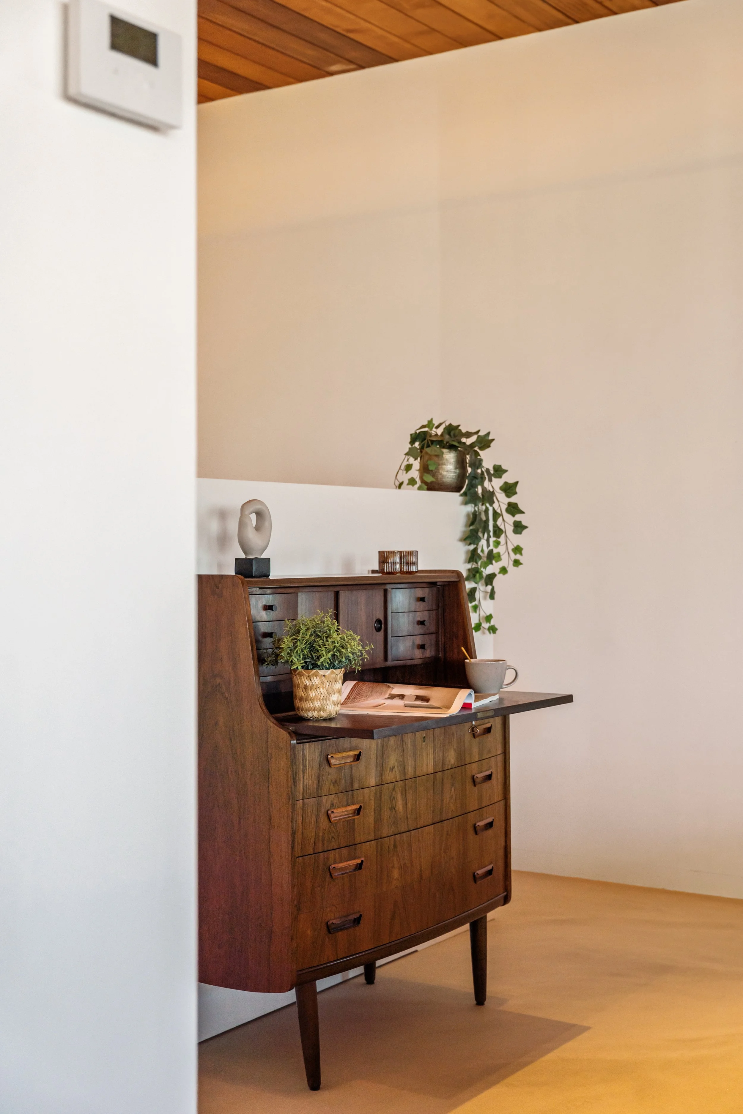 Een houten buffetkast met meerdere laden en een donker tafelblad, versierd met een plantenbak en een mok, in een kamer met witte muren en een houten plafond.