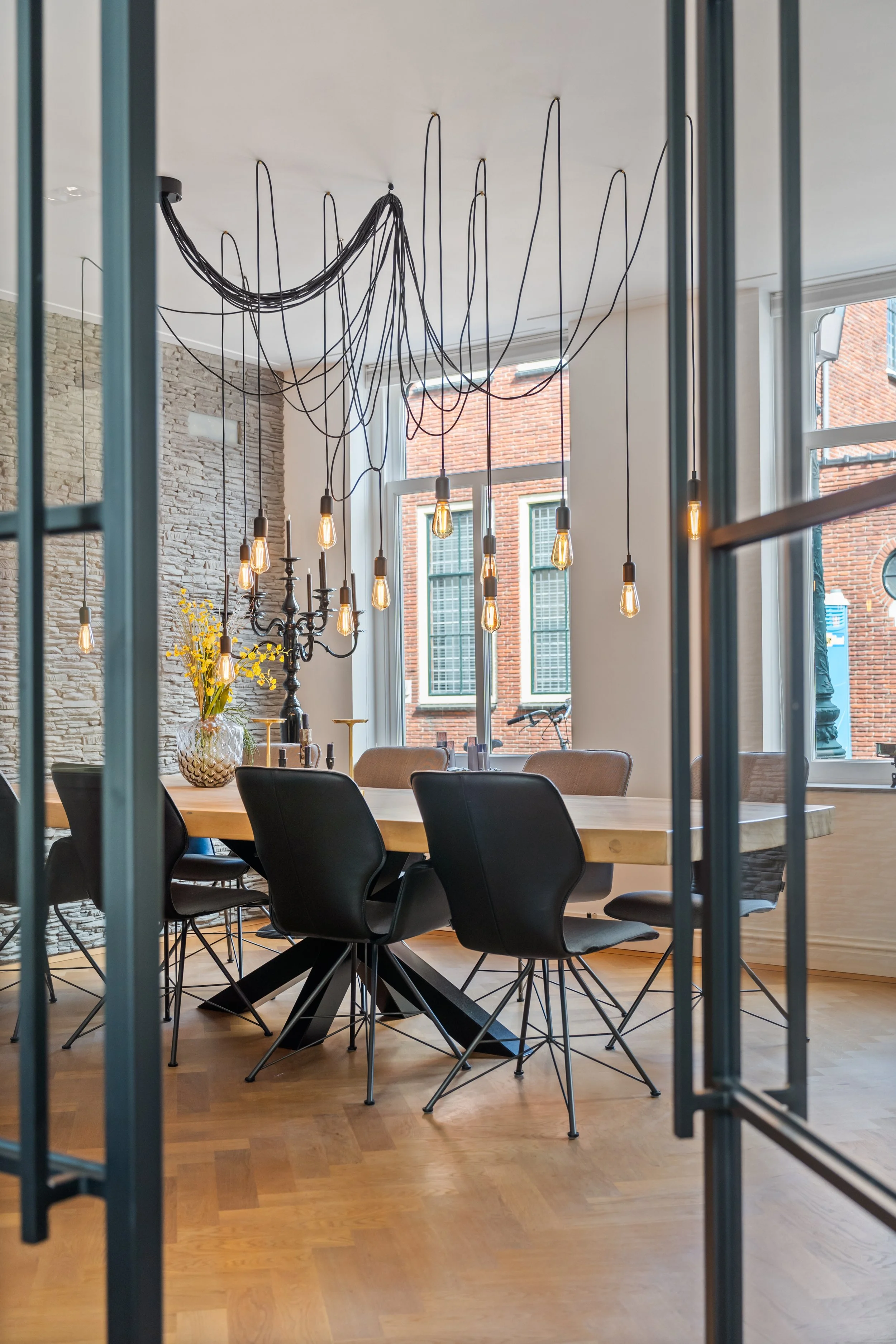 Interieur van een modern restaurant met een grote houten eettafel, zwarte stoelen, hanging gloeilampen en grote ramen met uitzicht op een bakstenen gebouw.