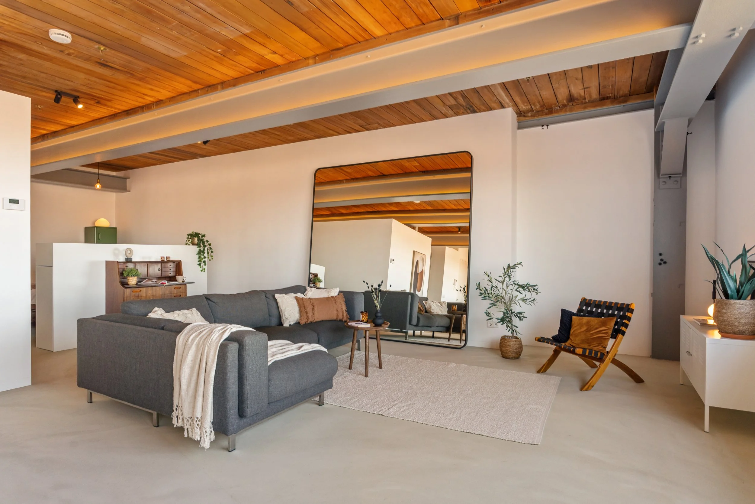 Woonkamer met houten plafond, grote spiegel, grijze bank, planten, en een gele en zwarte houten stoel.