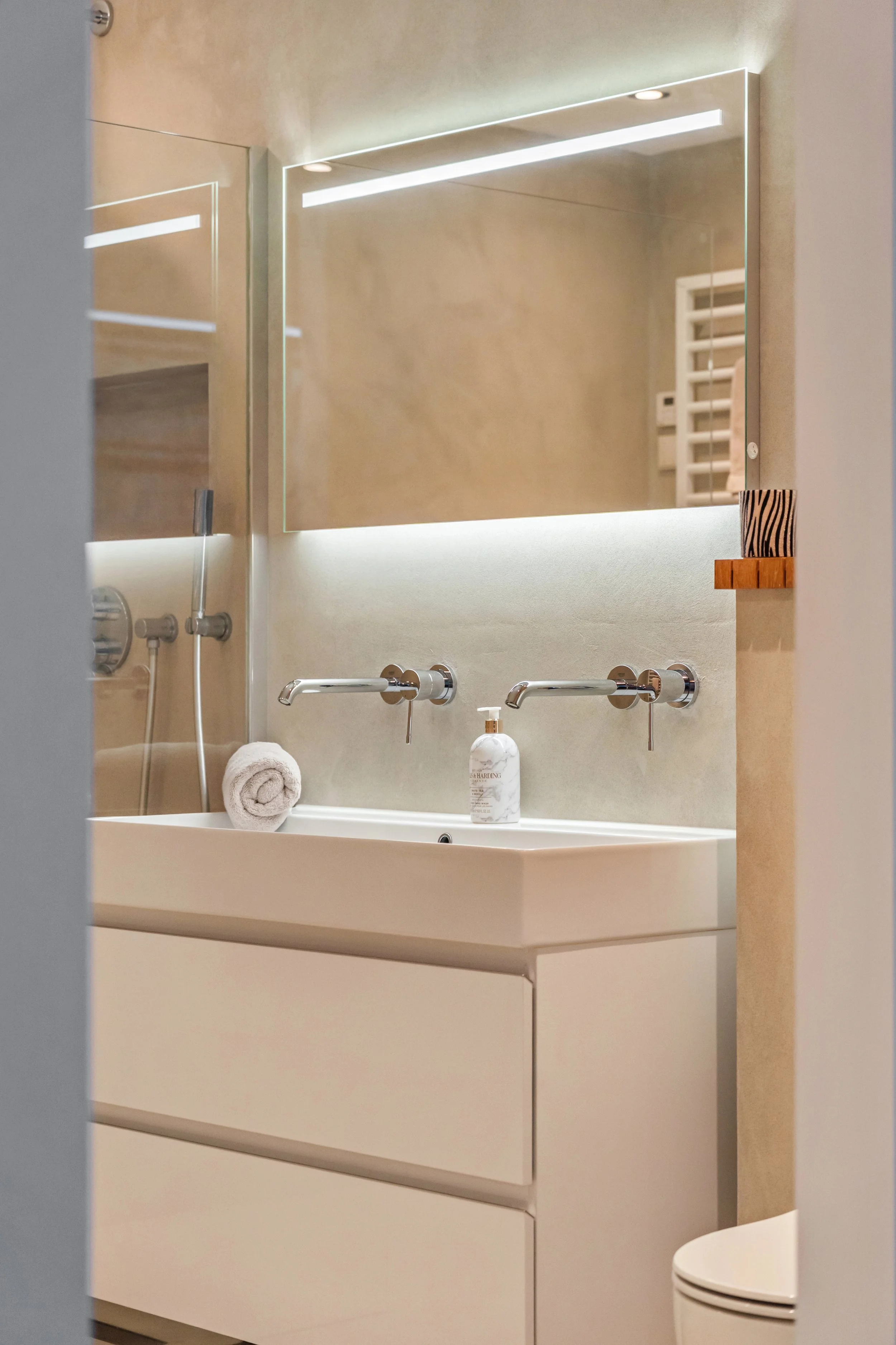 Moderne badkamer met wit wastafelmeubel, spiegel met ingebouwde verlichting, handdoek gerold, zeepfles en een toilet op de rechterkant.