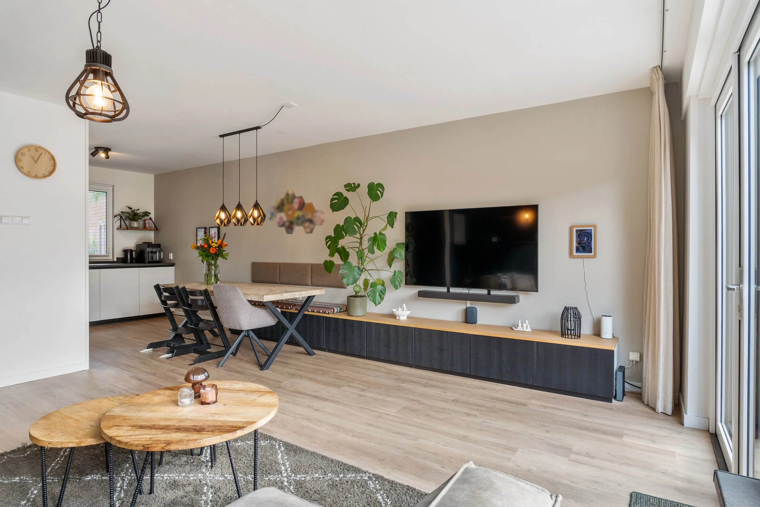 Een woon- of eetkamer met een grote flatscreen-tv aan de muur, een houten salontafel met decoraties, een eetkamertafel met stoelen, planten, en een keuken op de achtergrond. Grote ramen met gordijnen laten natuurlijk licht binnen.