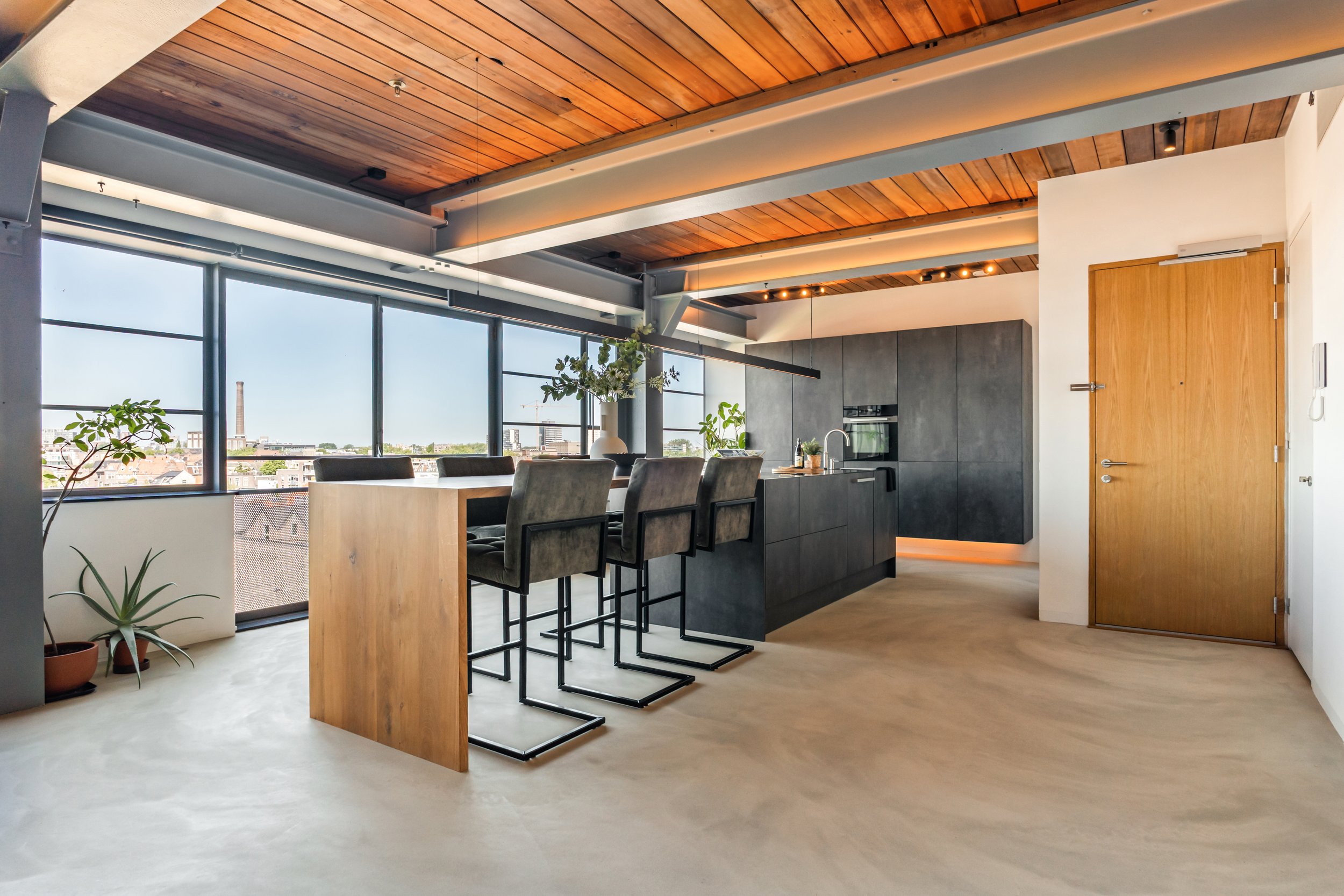 Moderne keuken en eethoek met houten plafond en grote ramen, uitzicht op stad met fabrieksschoorsteen, planten in potten, centraal eiland met barstoelen.