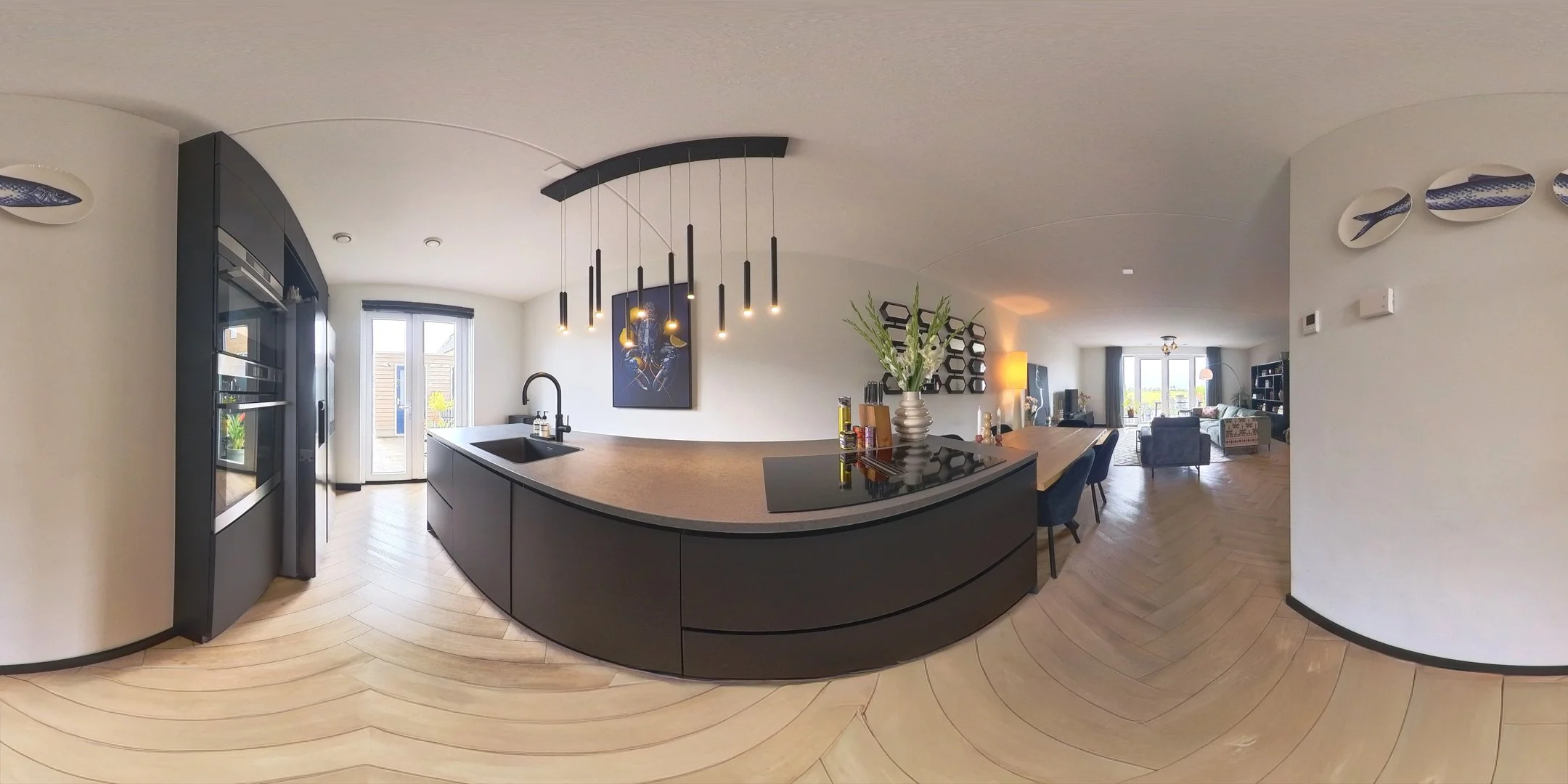 Een moderne keuken met een u-vormig aanrechtblad, hangende lampen, een zwart fornuis met een bloemenvaas, en een overzicht van de woonkamer met meubels en grote ramen.