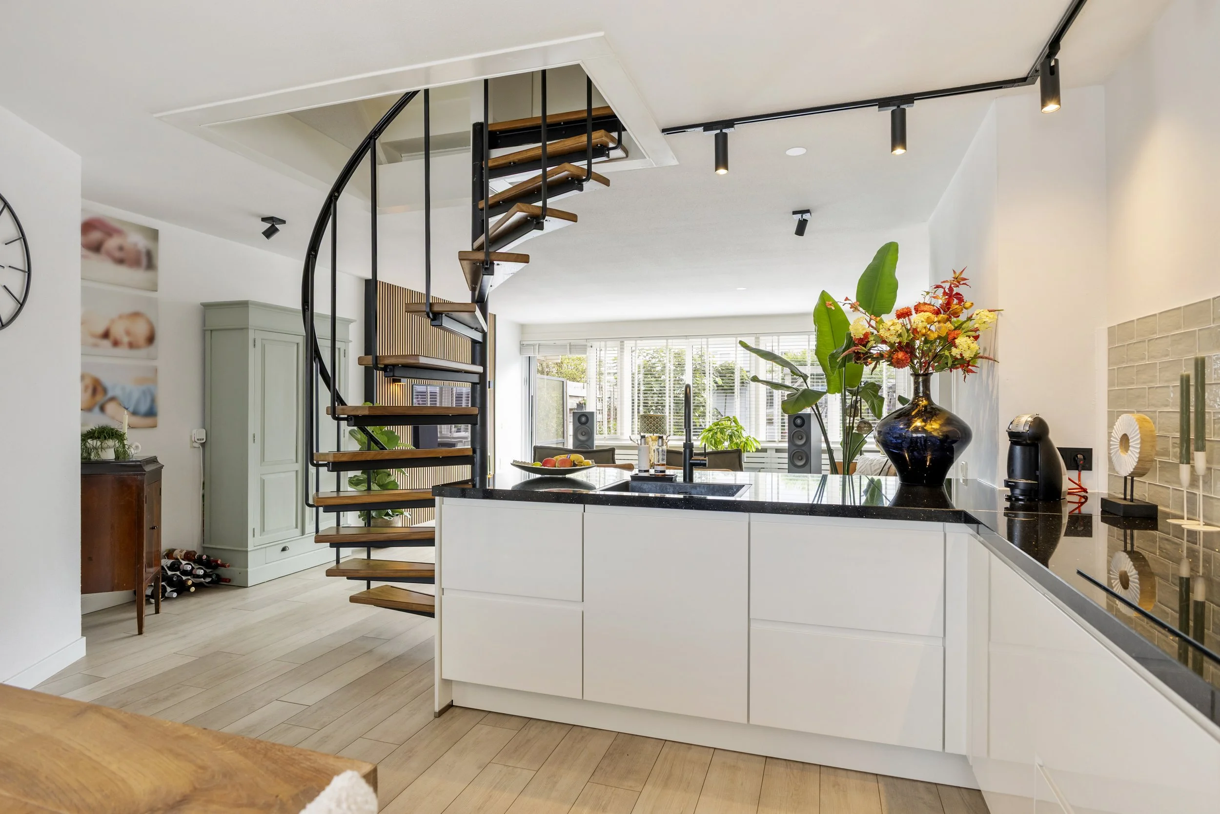 Moderne open keuken met een zwart granieten werkblad, grote bloemenvaas, eettafel met fruit, keukenappara­tuur en apparatuur, grote ramen met luiken, een groene kast en spinnende lampen.