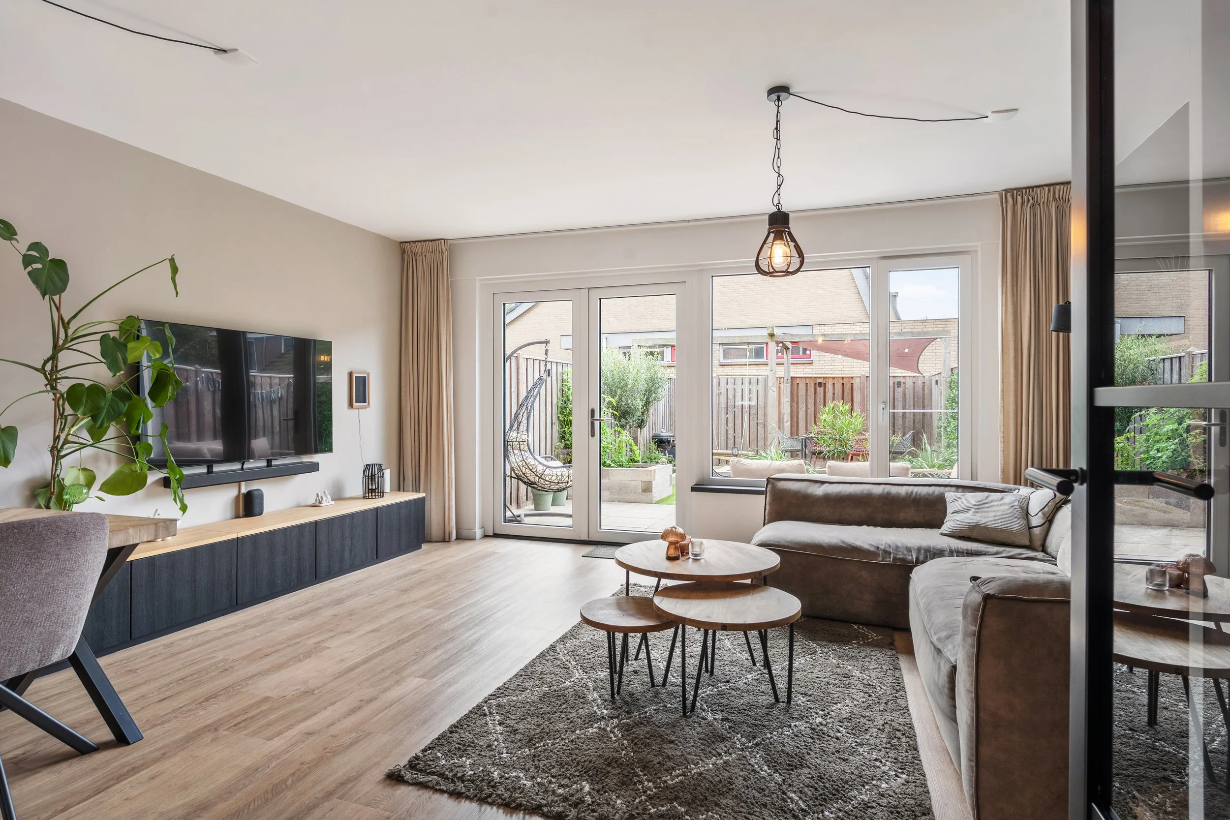 Een woonkamer met grote ramen en een deur die uitkijken op een tuin, met een bank, salontafels en een plant.