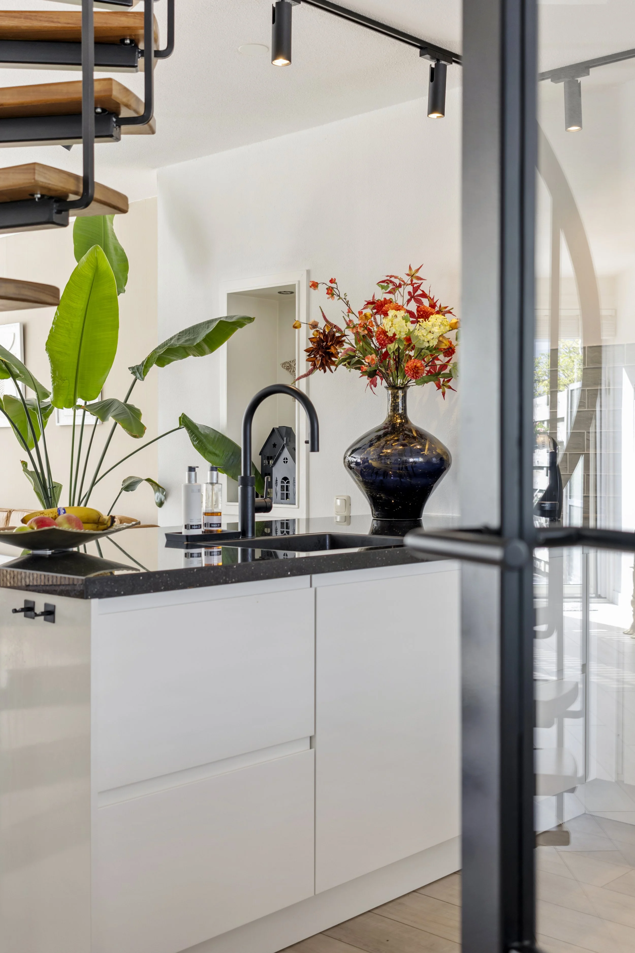 Een moderne keuken met witte kasten, een zwart granieten werkblad, een grote vaas met kleurrijke bloemen, groene planten, en een donkere muur met een groot raam.