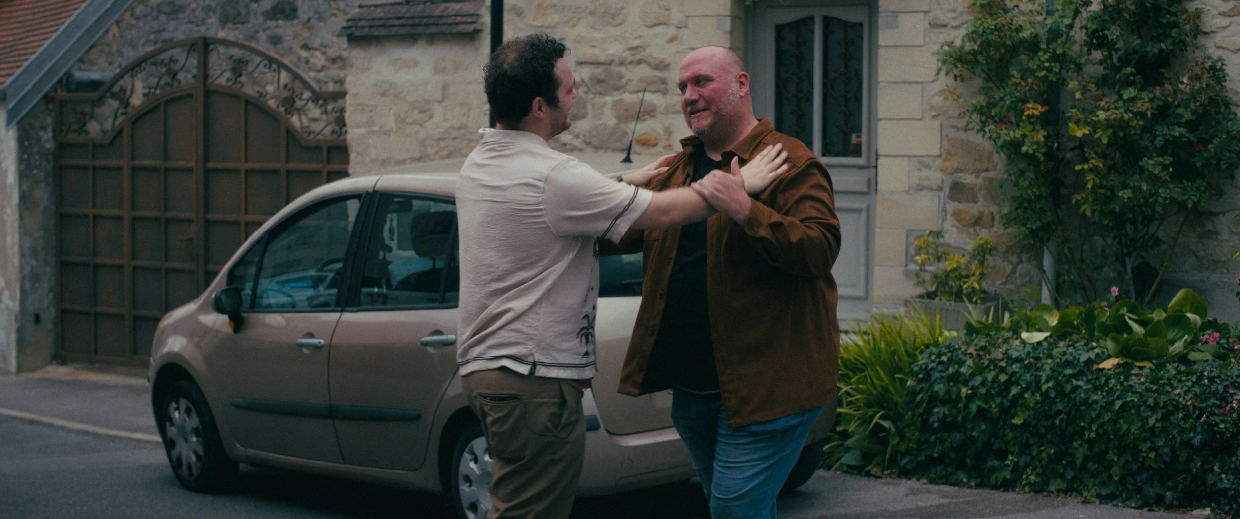 Two men are arguing outside in front of a house, with one grabbing the other by the collar and the other holding his shoulders. A small beige car is parked nearby, and there are plants and a stone wall in the background.