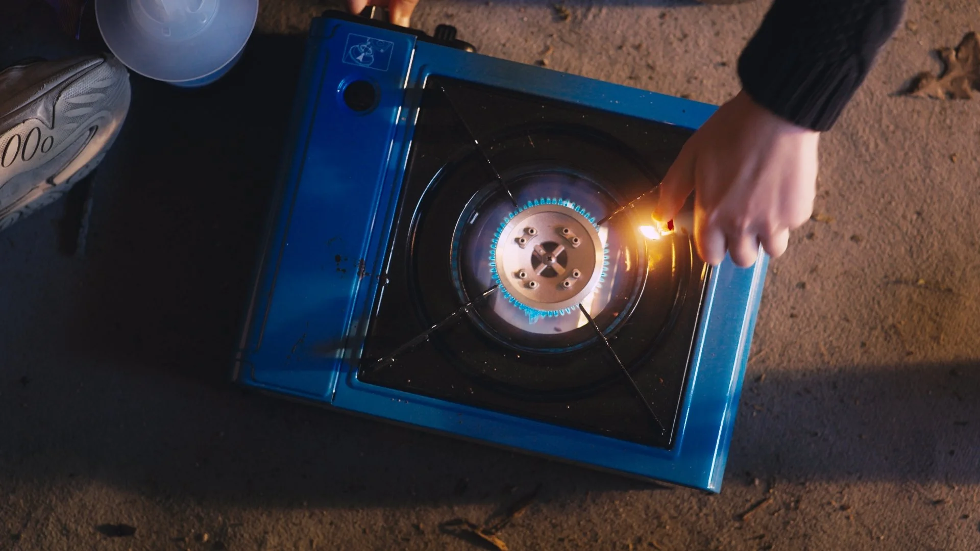 A person using a blue portable stove with a visible flame on a sandy ground, wearing a black jacket and holding a lighter near the stove.