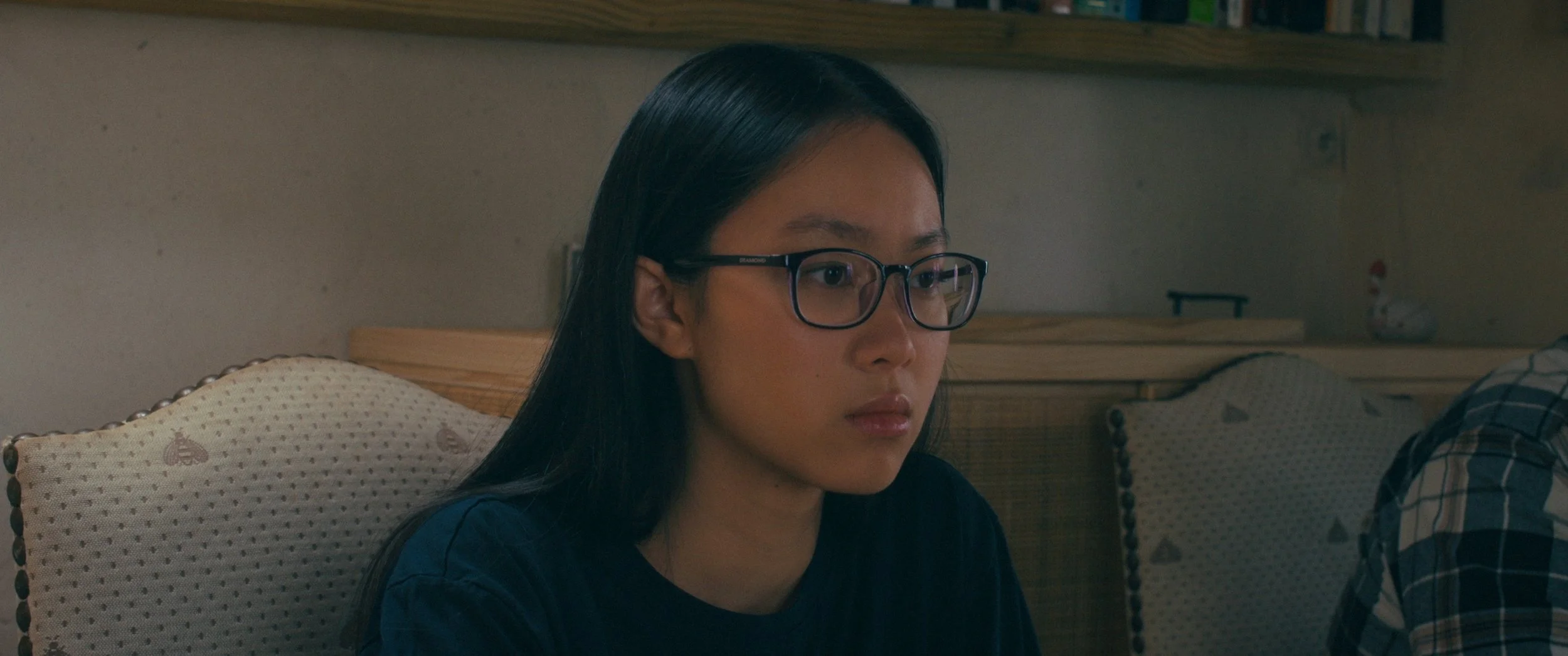 A young woman with long black hair and glasses sitting on a couch, looking down with a serious expression.