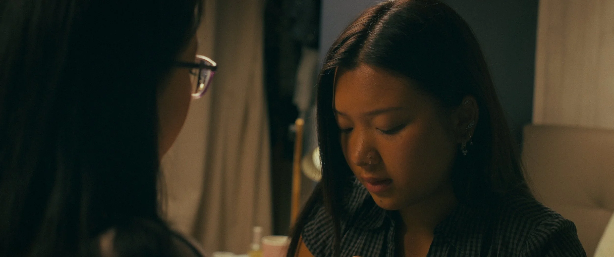 A young woman with long dark hair and a nose piercing, appearing emotional, is being comforted or spoken to by another person whose face is not visible in a dimly lit room.