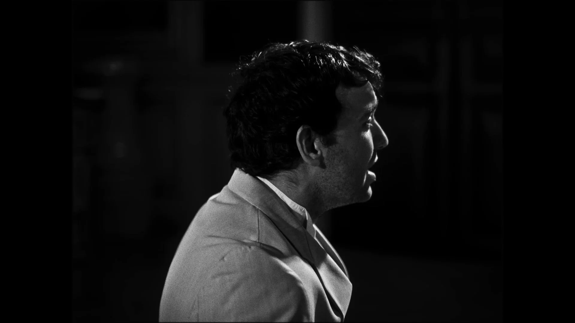 Black and white side profile of a man with dark, wavy hair, wearing a light-colored jacket, in a dark room.