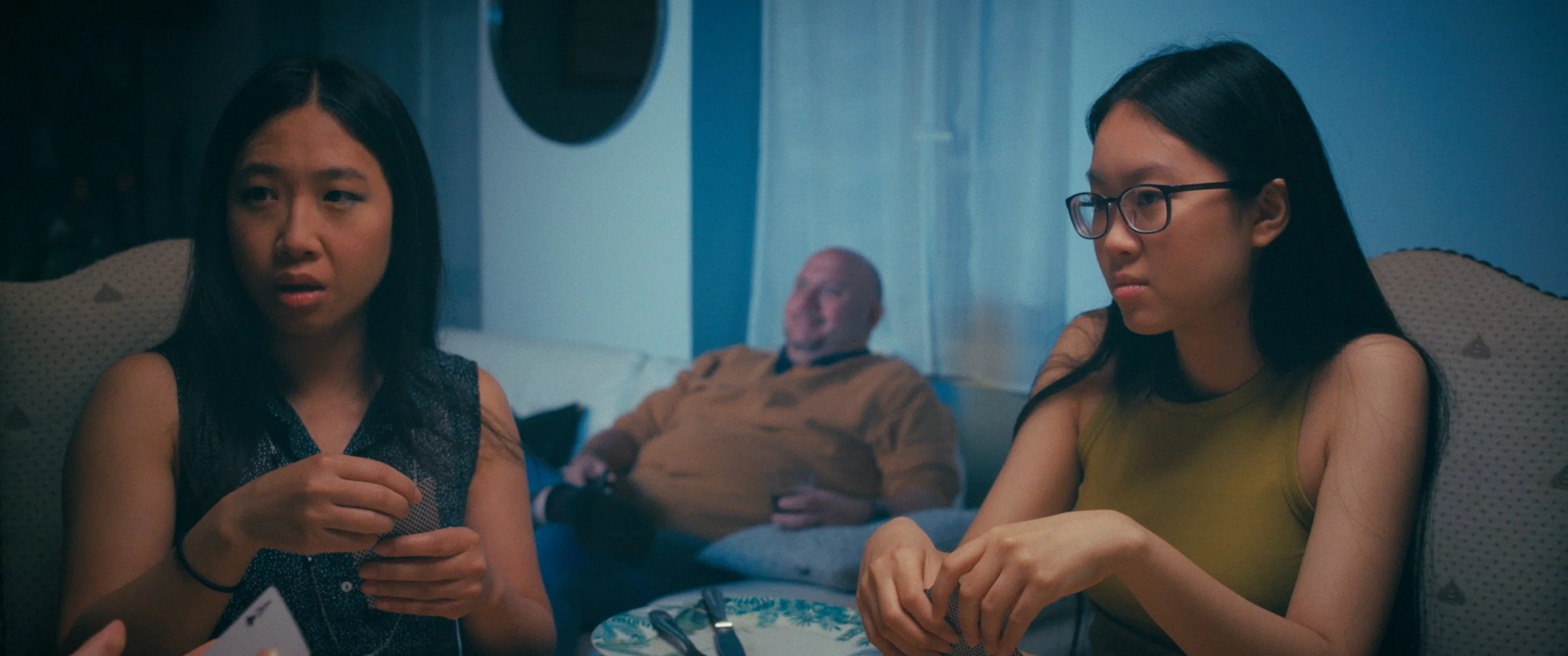 Two women sit at a table, happy, while a man in the background is watching tv.