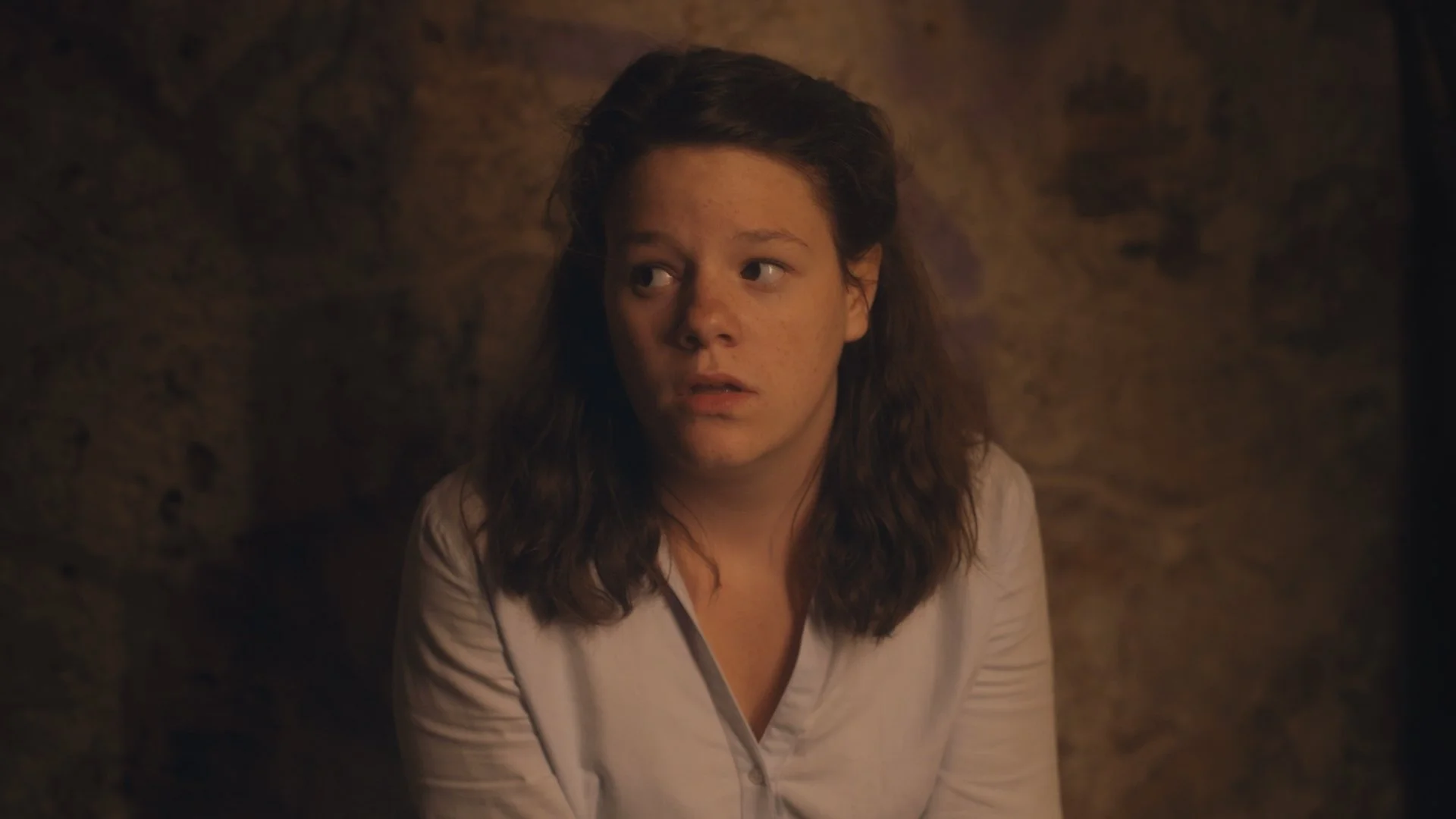 A woman with brown hair and wearing a light blue top, sitting against a rough stone wall, looking to her left with a serious expression.