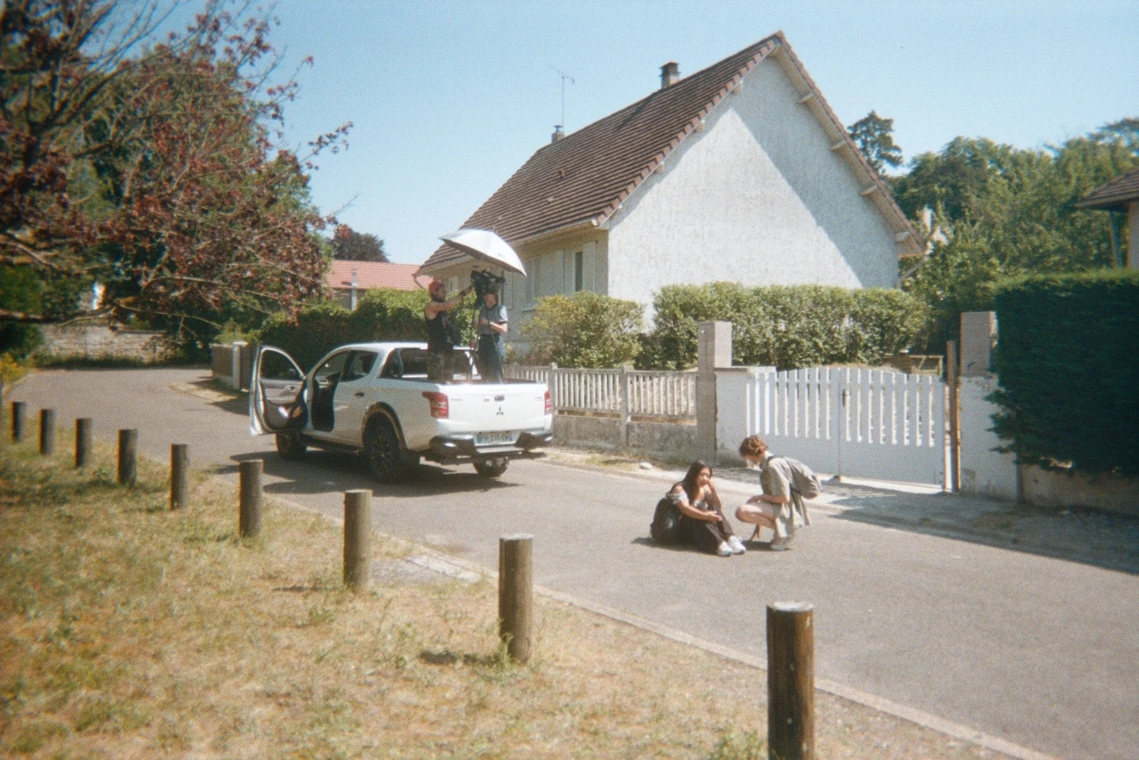 A residential street scene featuring a white house with a pitched roof, a parked silver car with an open door, and two people sitting on the sidewalk engaged in conversation, with trees and another house in the background.