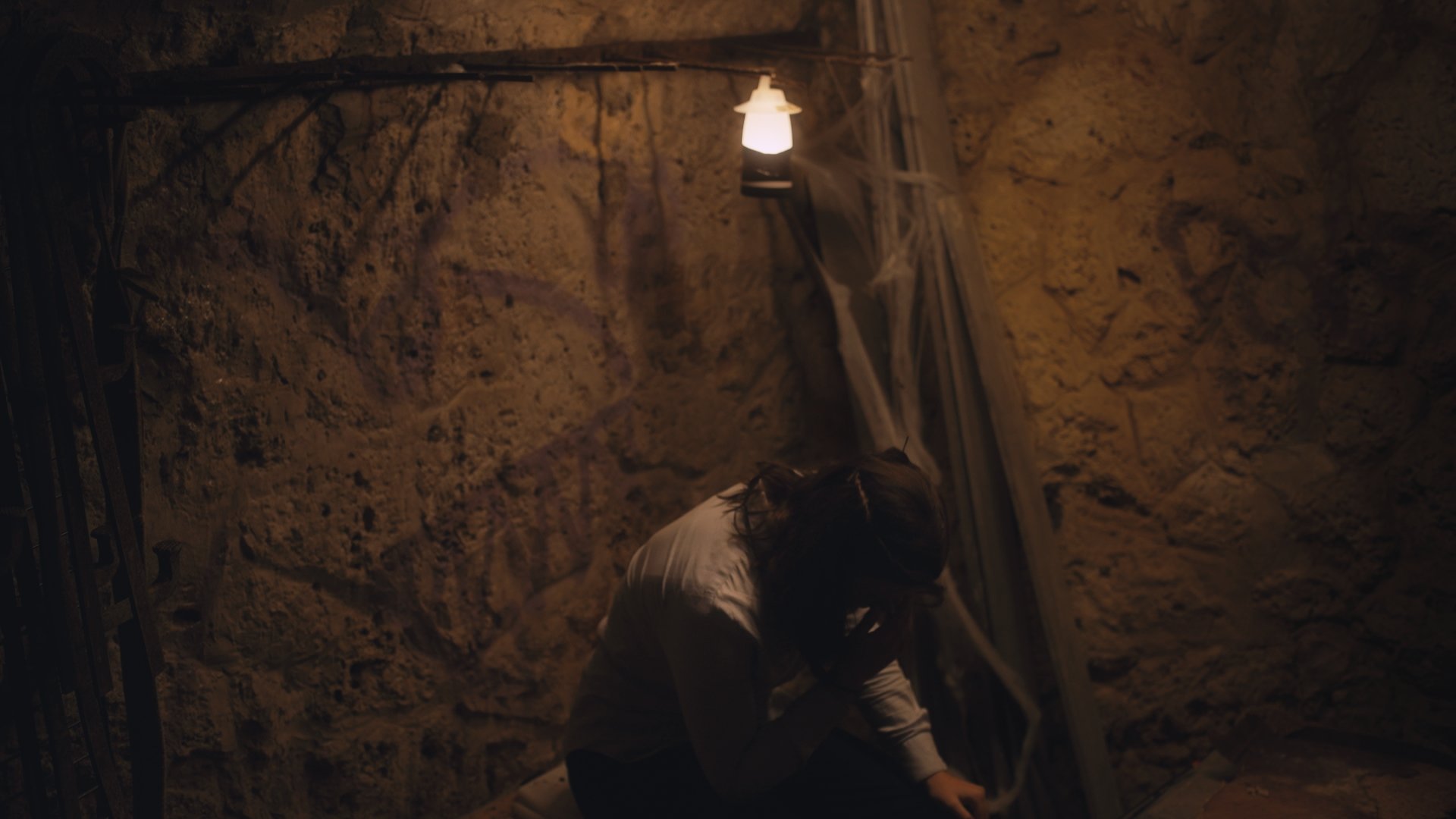 A person with long hair wearing a white shirt crouching down in a dimly lit stone or dirt tunnel with a single light hanging from above.
