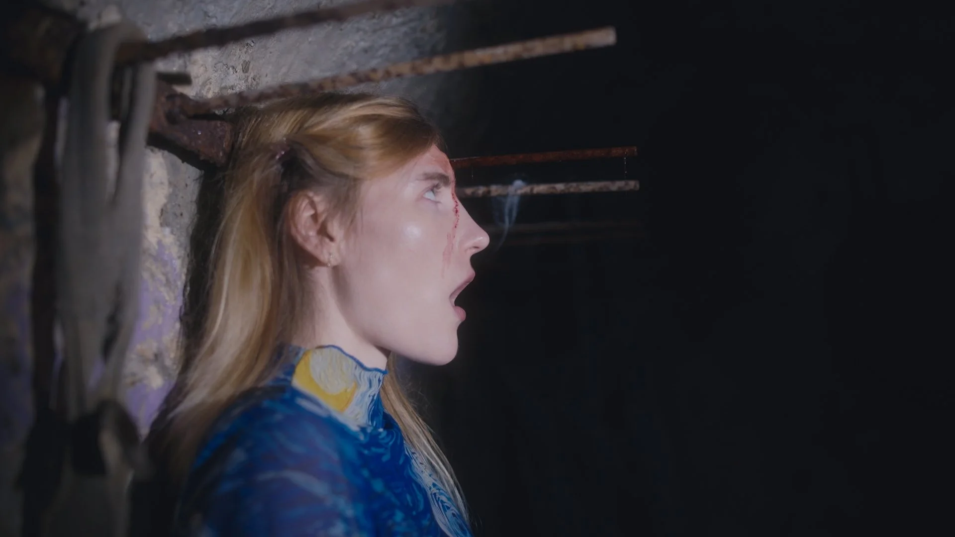 A woman with long blonde hair and painted blue top is shown with her mouth open with a rusty metal rod passing through her head in profil.