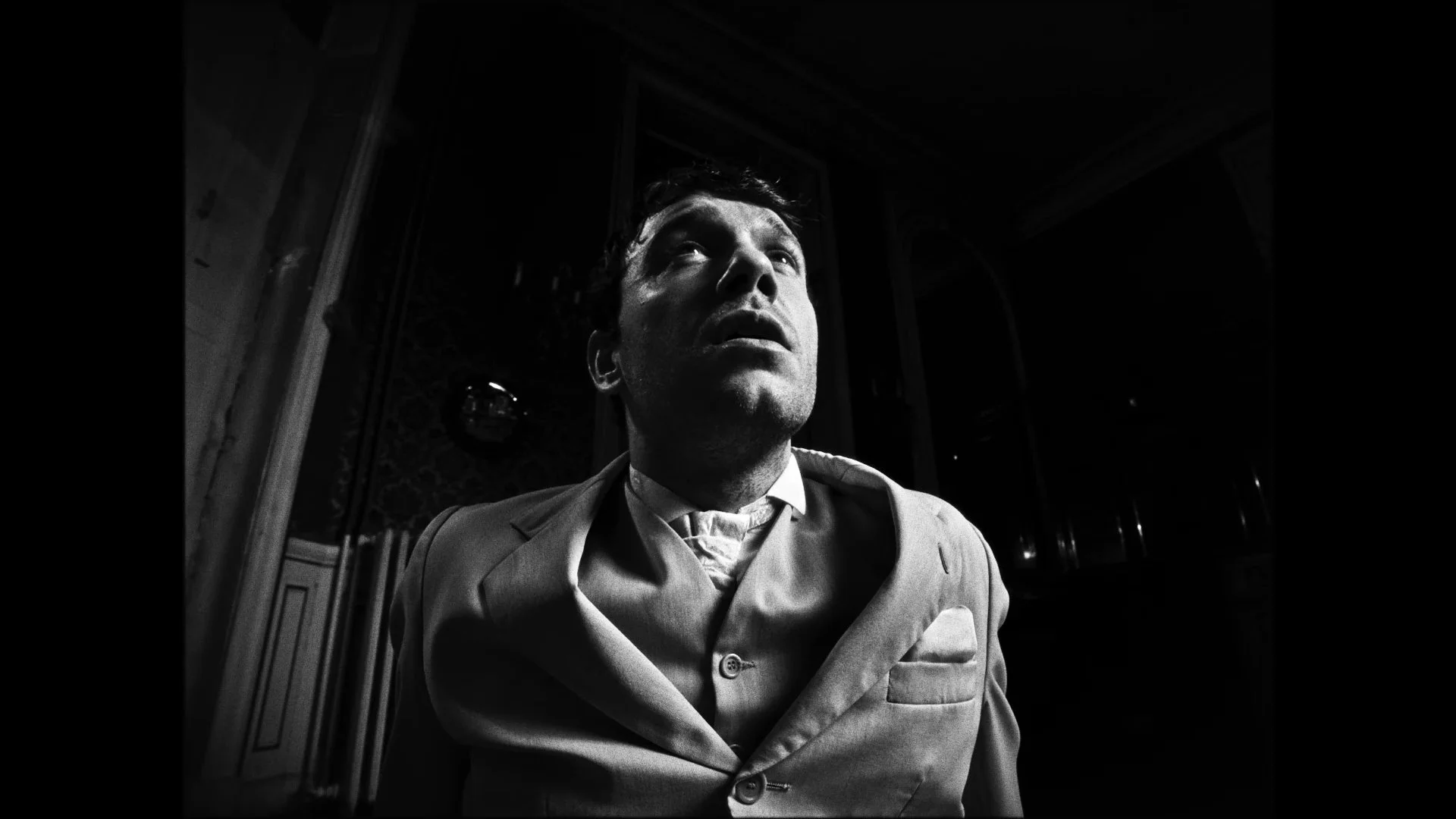 Black and white photo of a man in a suit looking upward with a serious expression, in a dimly lit room.