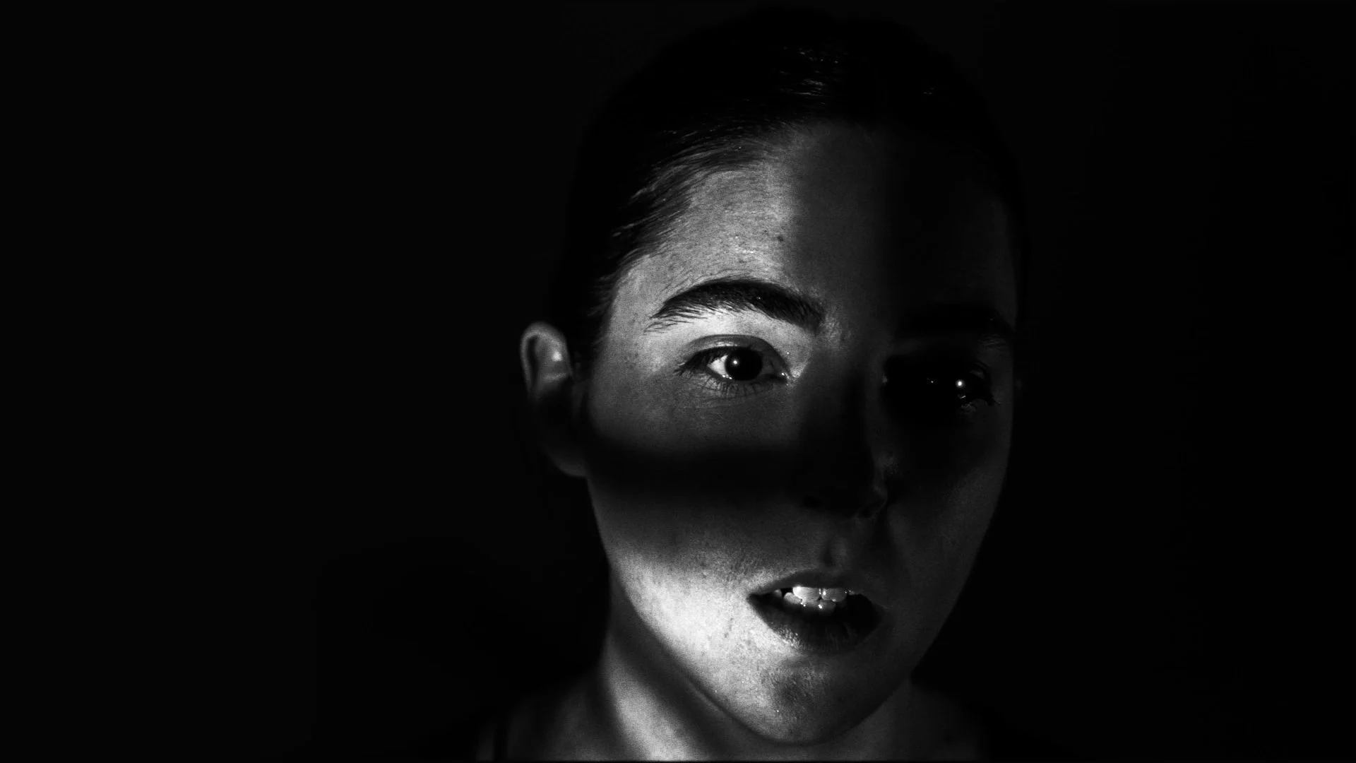 A black and white photo of a woman's face partially shadowed, with straight hair, high cheekbones, and expressive eyes.
