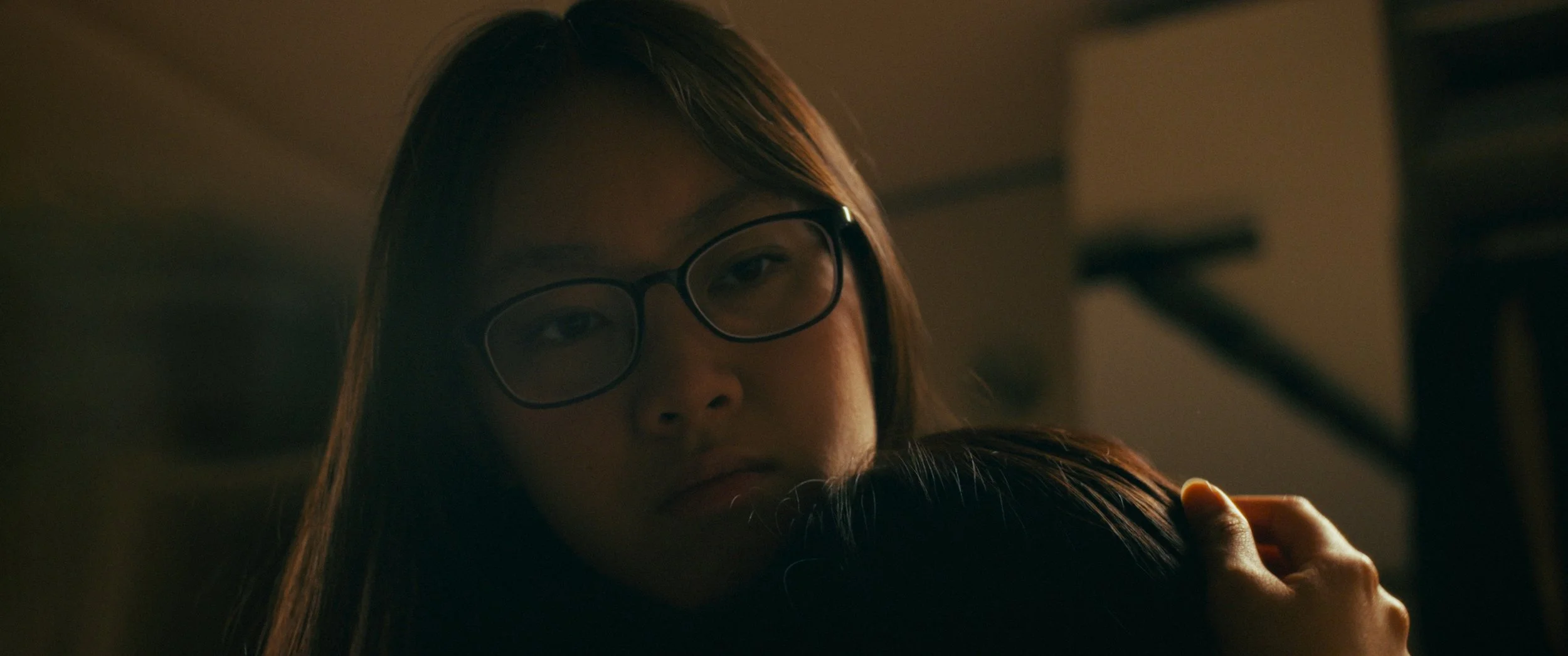 A woman with glasses and long hair gently hugging someone, with a dimly lit background.