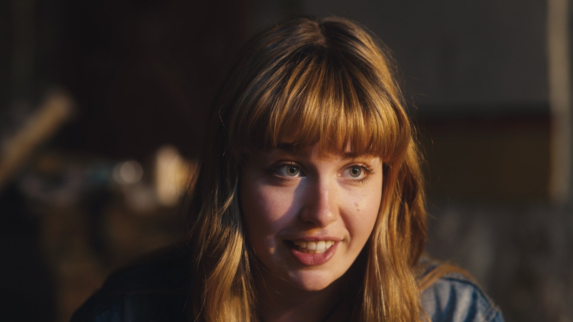 A young woman with light skin, long blond hair with bangs, and bright eyes, smiling and looking slightly upward, in a warmly lit environment.