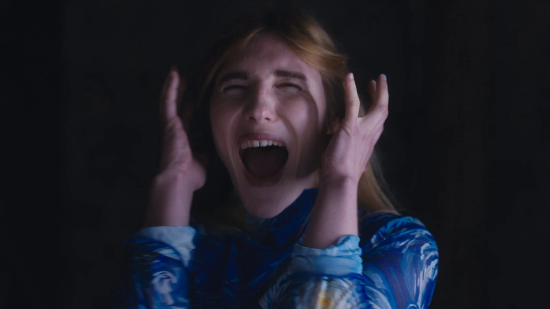 A woman with blond hair and a blue patterned shirt is yelling with her hands over her ears in a dark room.