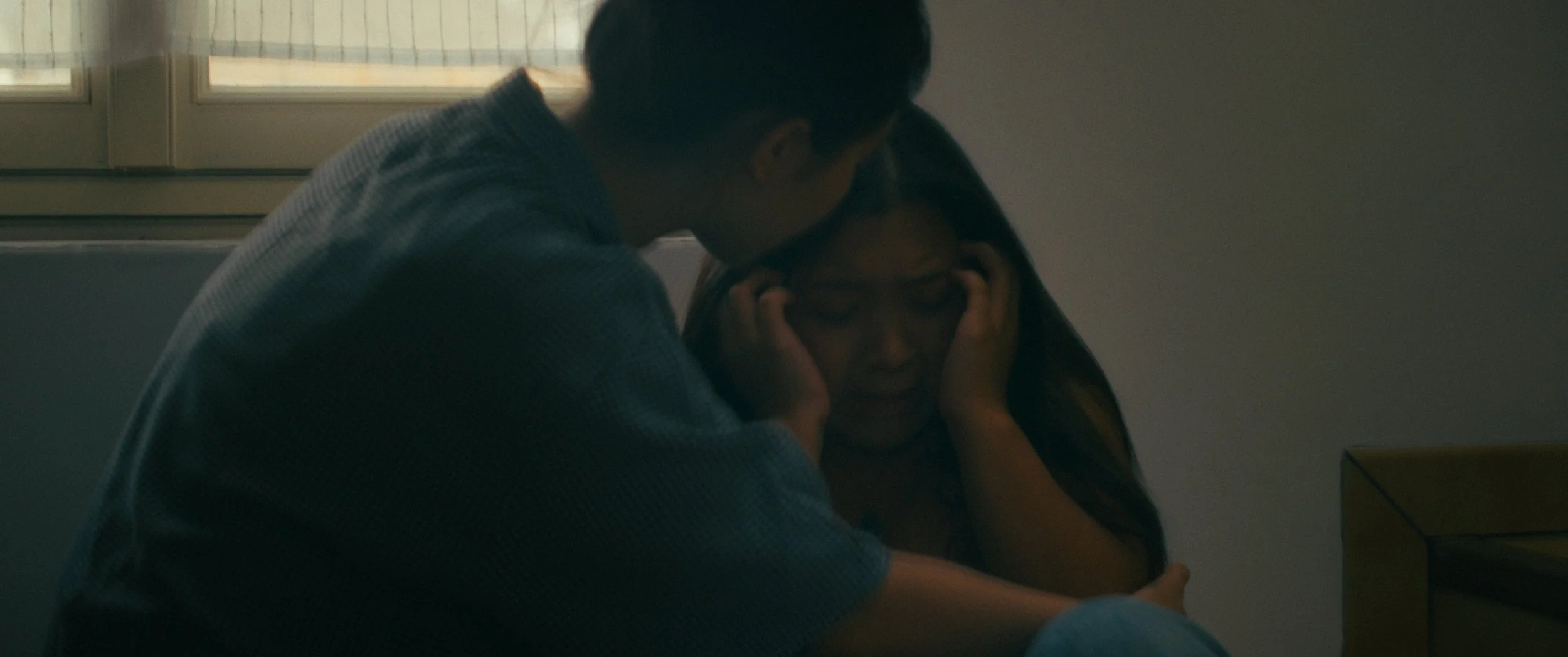 A young man comfortingly holds a young woman, who appears upset, in a dimly lit room near a window.