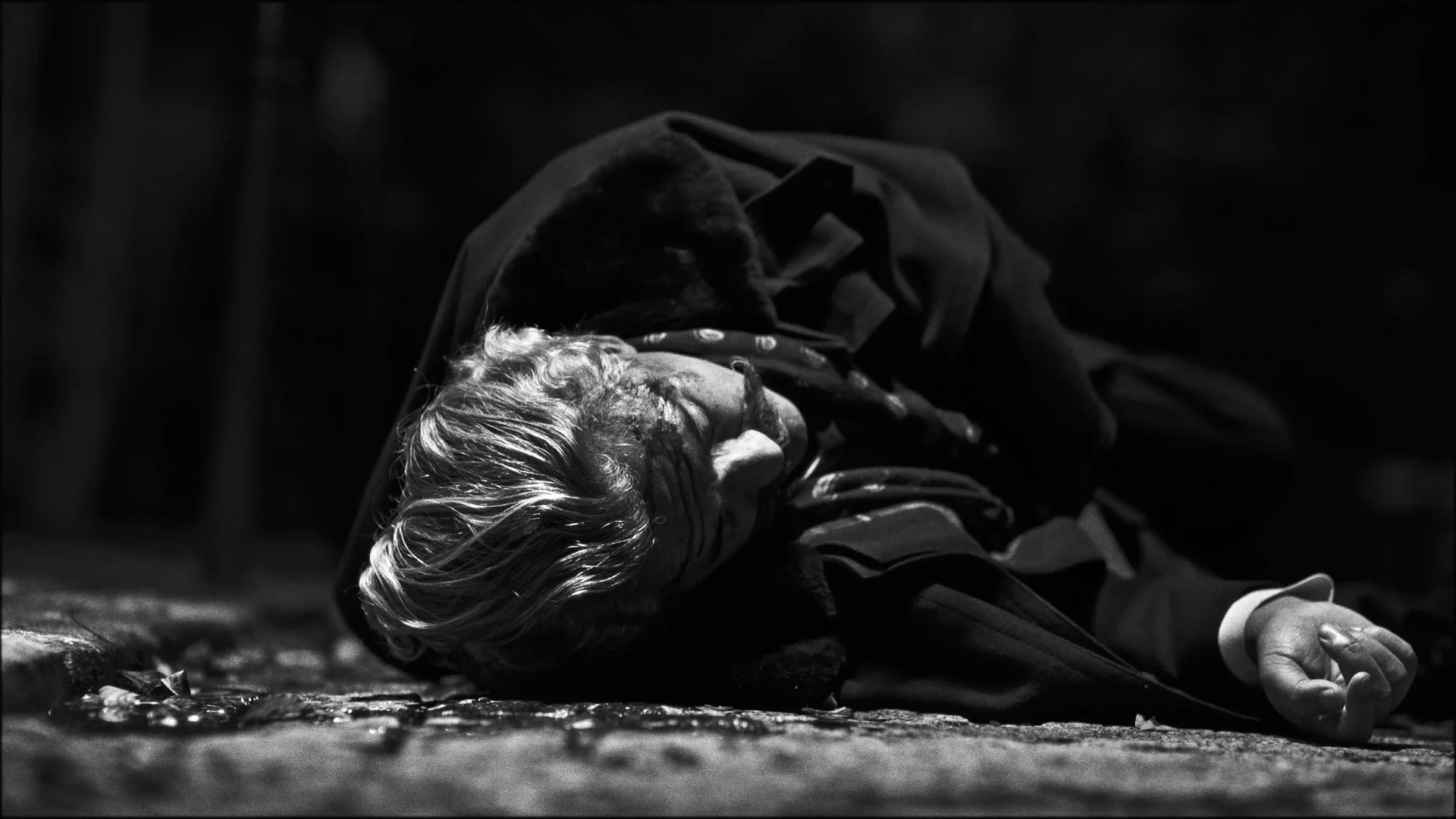 A person with light-colored hair lying on the ground, appearing unconscious or injured, with a dark environment around them.