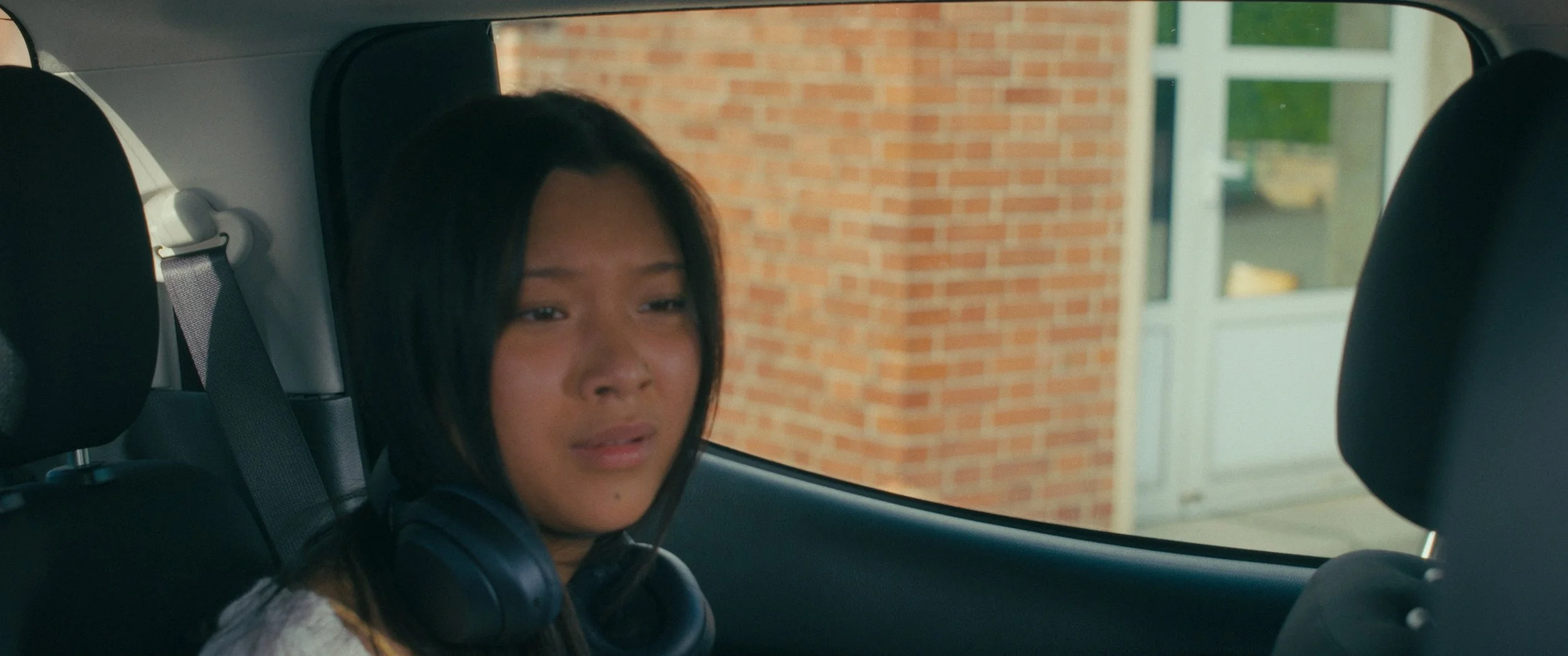 A young woman with long black hair wearing headphones, sitting in the backseat of a car, looking out the window with a neutral expression.