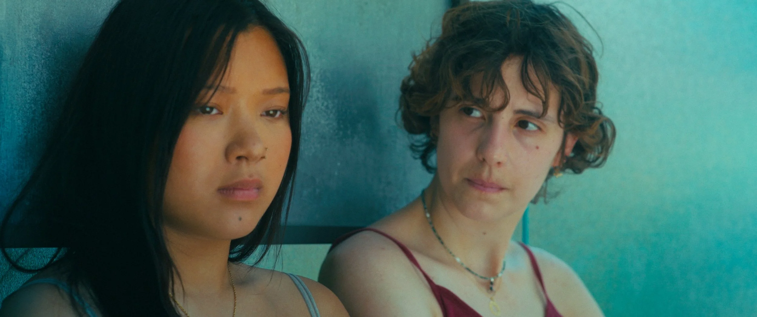 Two young women sitting side by side, looking contemplative, at a busstop. One has long black hair and the other has short, curly brown hair. Both are wearing tank tops and necklaces.