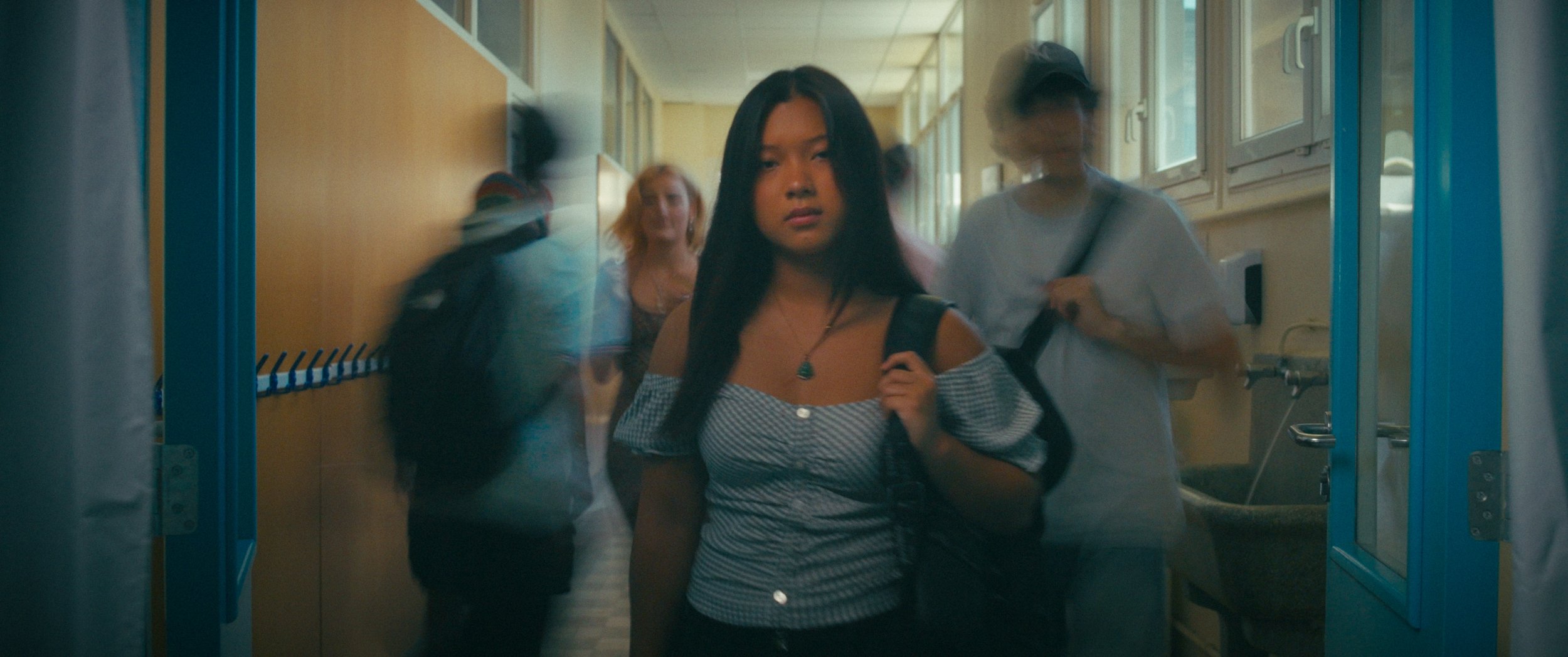 Teen girl with long black hair, wearing a striped off-shoulder top, carrying a backpack, walking through a school hallway while other students walk past her.
