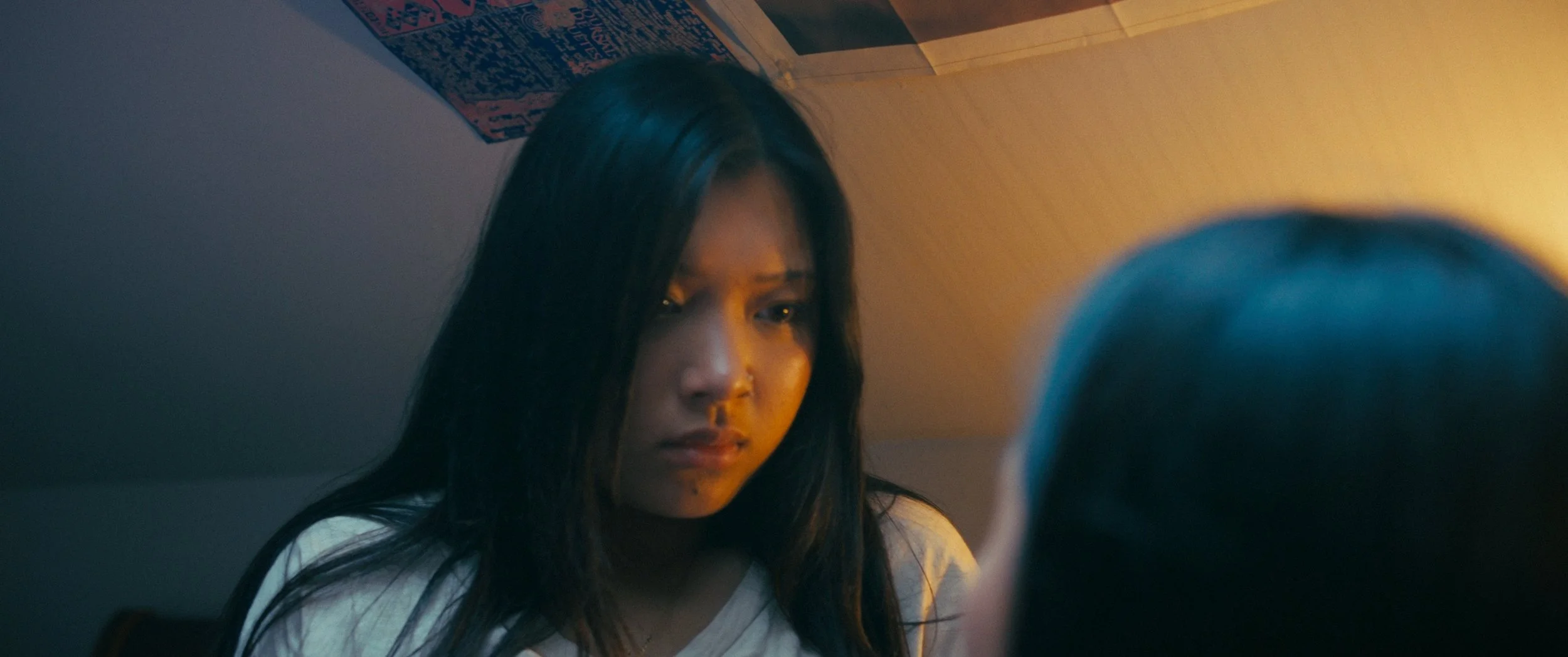 A young woman with long dark hair looks down, illuminated by warm lighting, with a poster on the wall behind her.