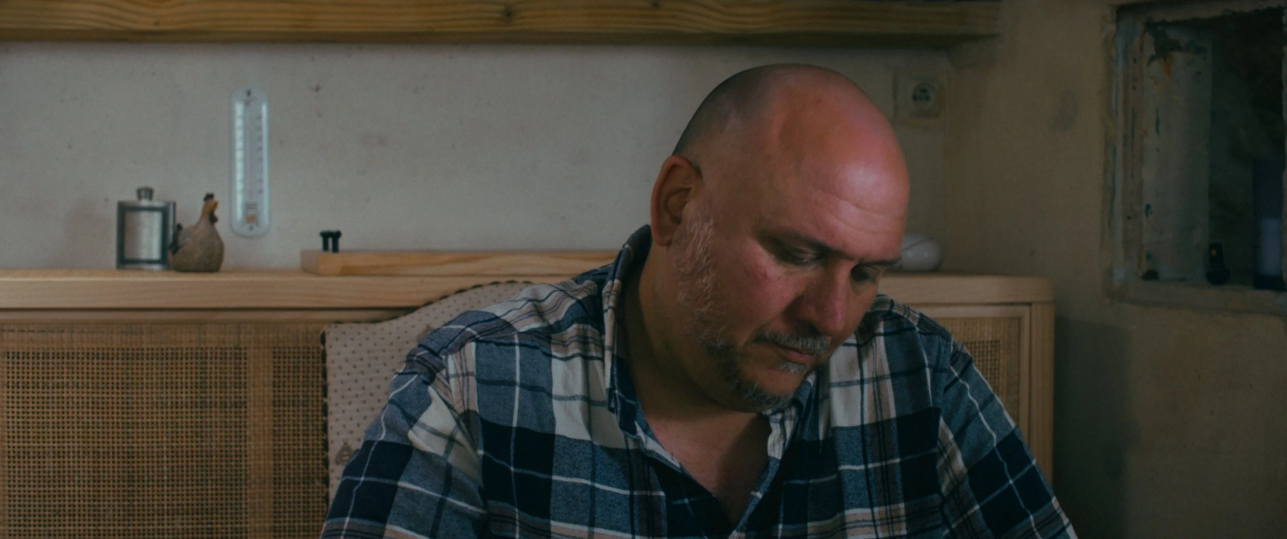 A bald man with a beard is sitting at a table, looking downward, in a room with a wooden shelf and a window in the background.