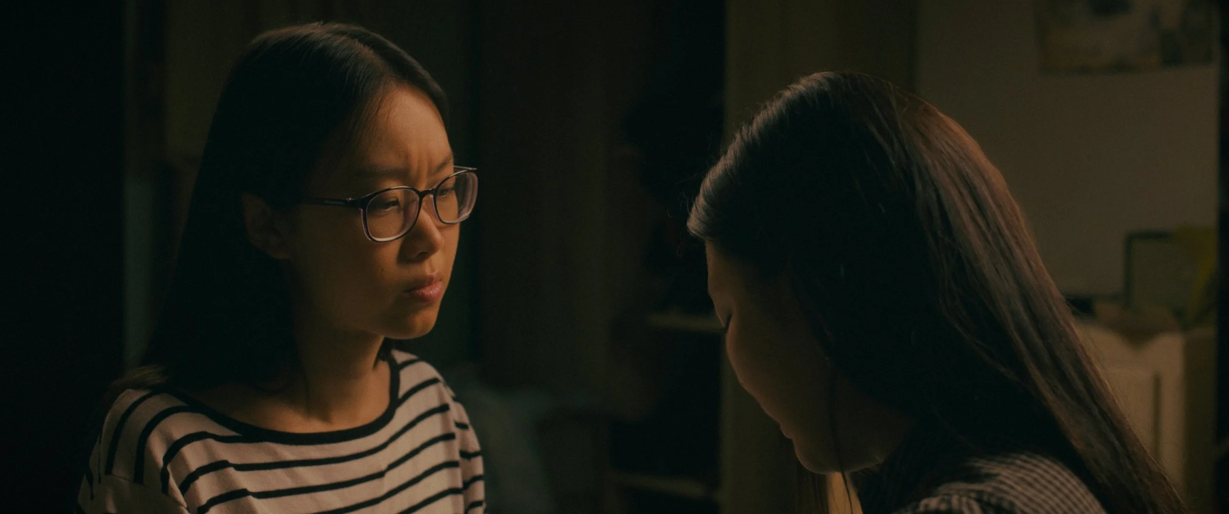 Two women face each other in a dimly lit room, with the woman on the left wearing glasses and a striped shirt appearing upset or concerned while the woman on the right has her head bowed.