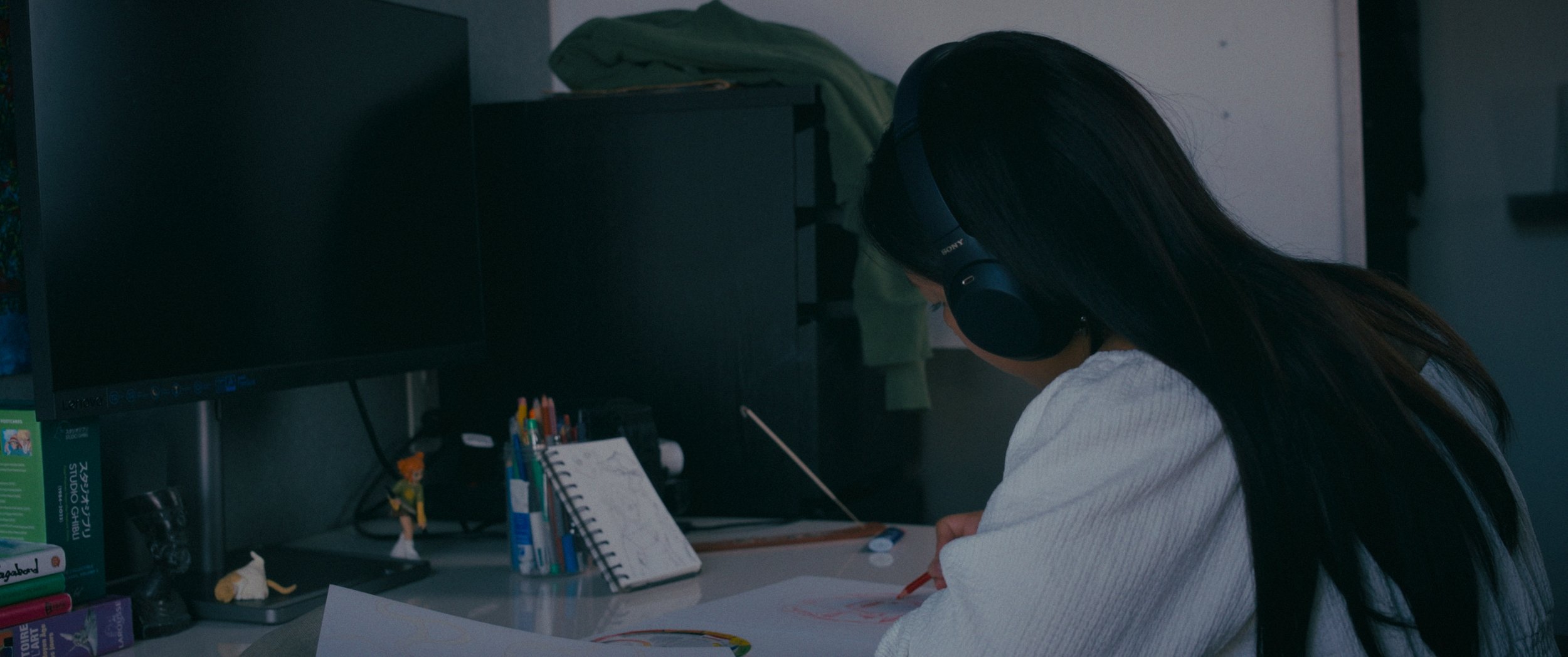 A young woman with long dark hair wearing black headphones and a white sweater, sitting at a desk, drawing on a sheet of paper with colored markers on a cluttered desk in front of a computer monitor.