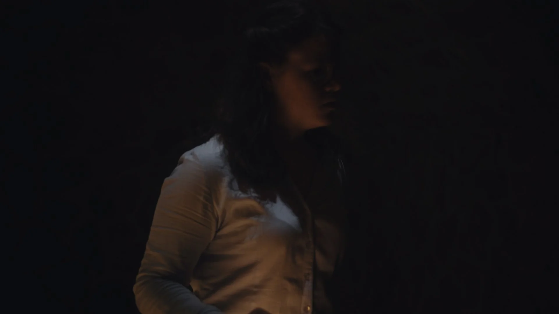 A woman with dark hair wearing a blue shirt, face turned to the side, in a dark environment.