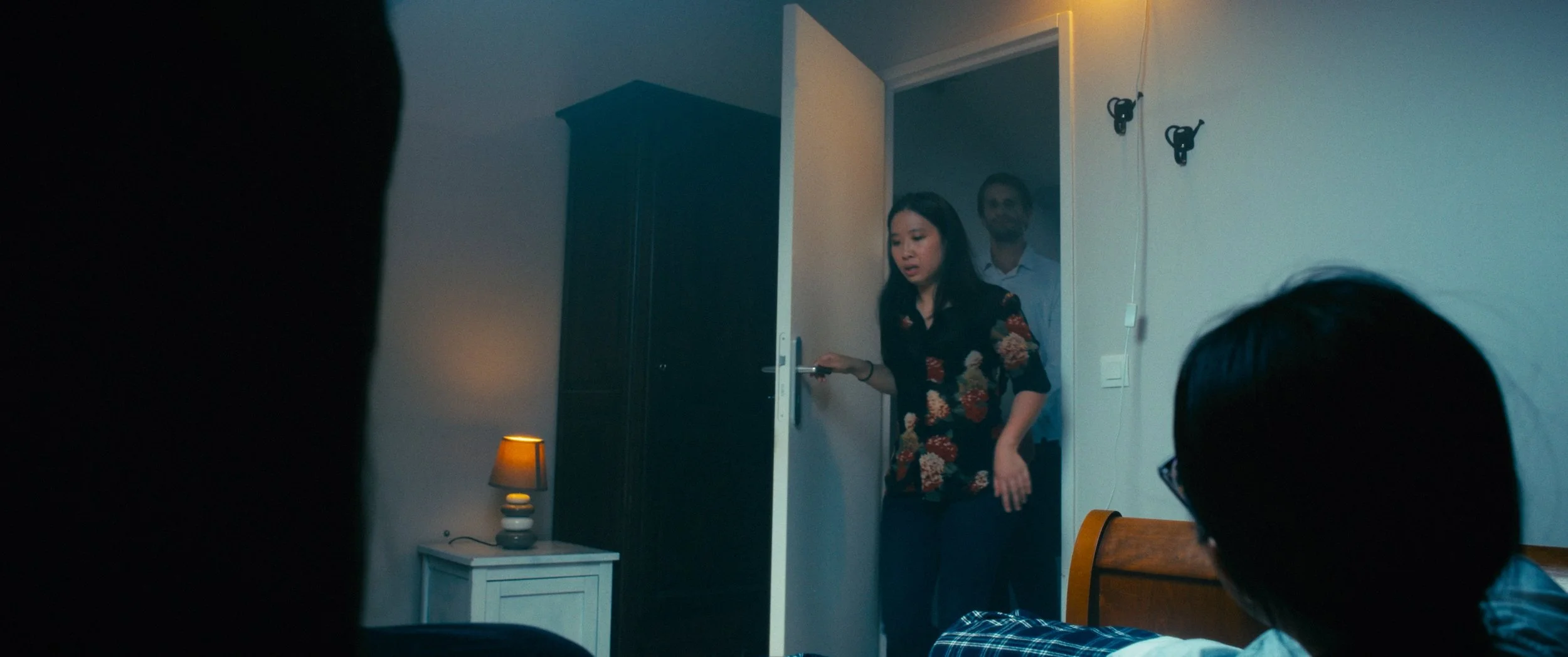 A woman in a floral shirt is entering a room with a door open, holding the handle, while a man stands behind her. In the foreground, a person with glasses is sitting on a bed, and parts of two other people are visible at the edges of the image. The r