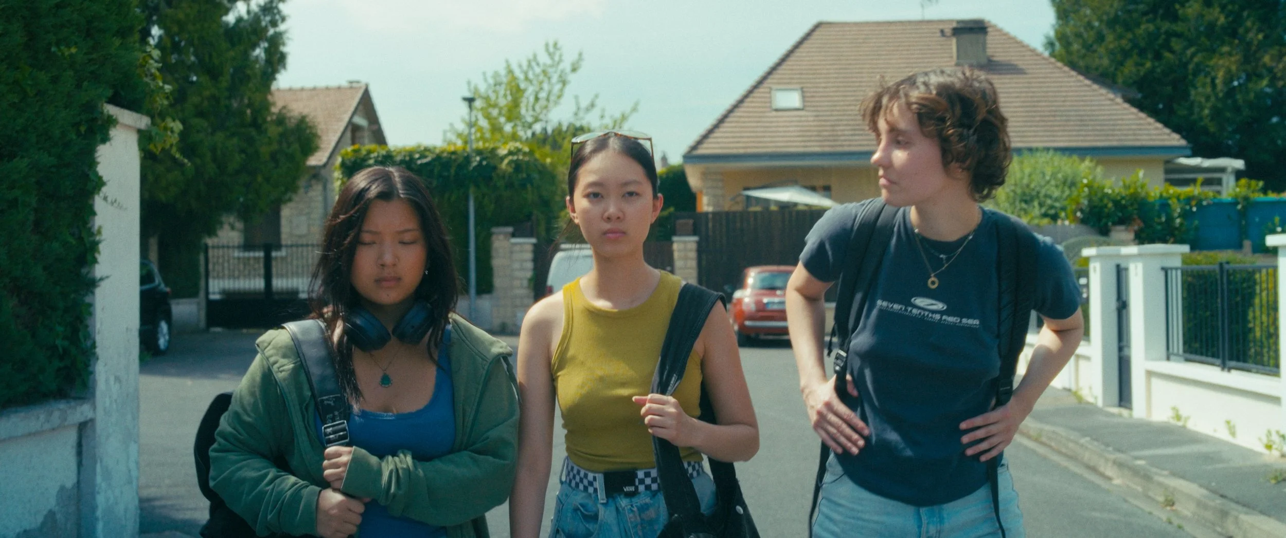 Three teenage girls walking on a suburban street, one with eyes closed, another looking directly ahead, and the third looking to the side.