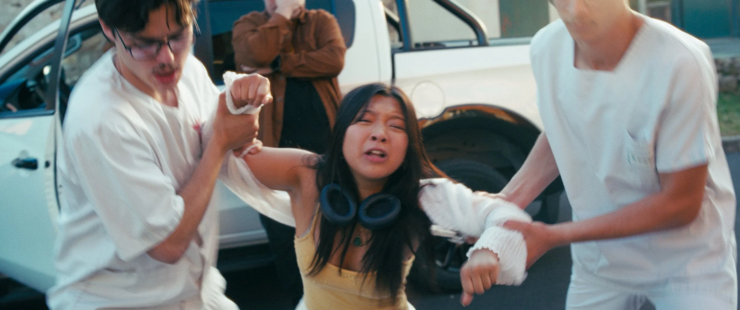 A woman in distress being assisted by two men, with a car in the background.