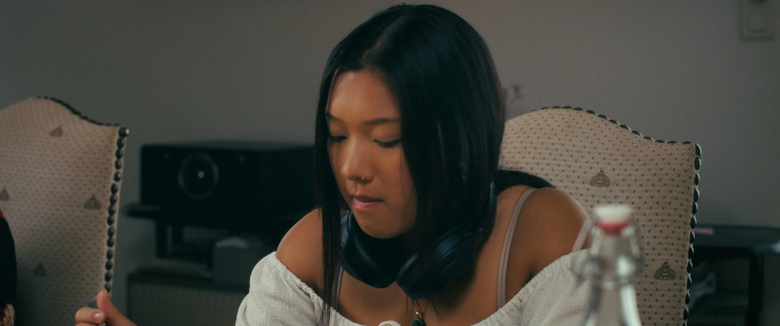 A woman with black hair sitting at a table in a room, wearing headphones around her neck, appearing focused or upset.