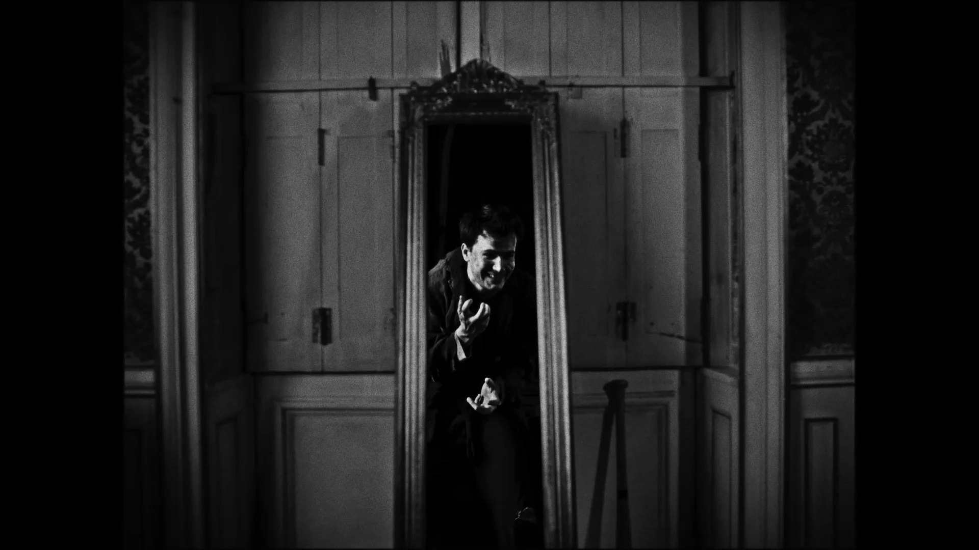 A man looking into a tall mirror in a room with wood-paneled walls, smiling and gesturing with his hand.