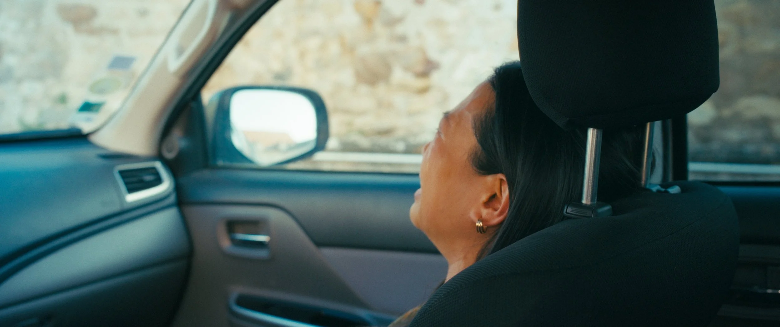 A woman with dark hair and earrings sitting in the passenger seat of a car, leaning back with her head resting against the headrest. The car's interior and a stone wall outside the window are visible.