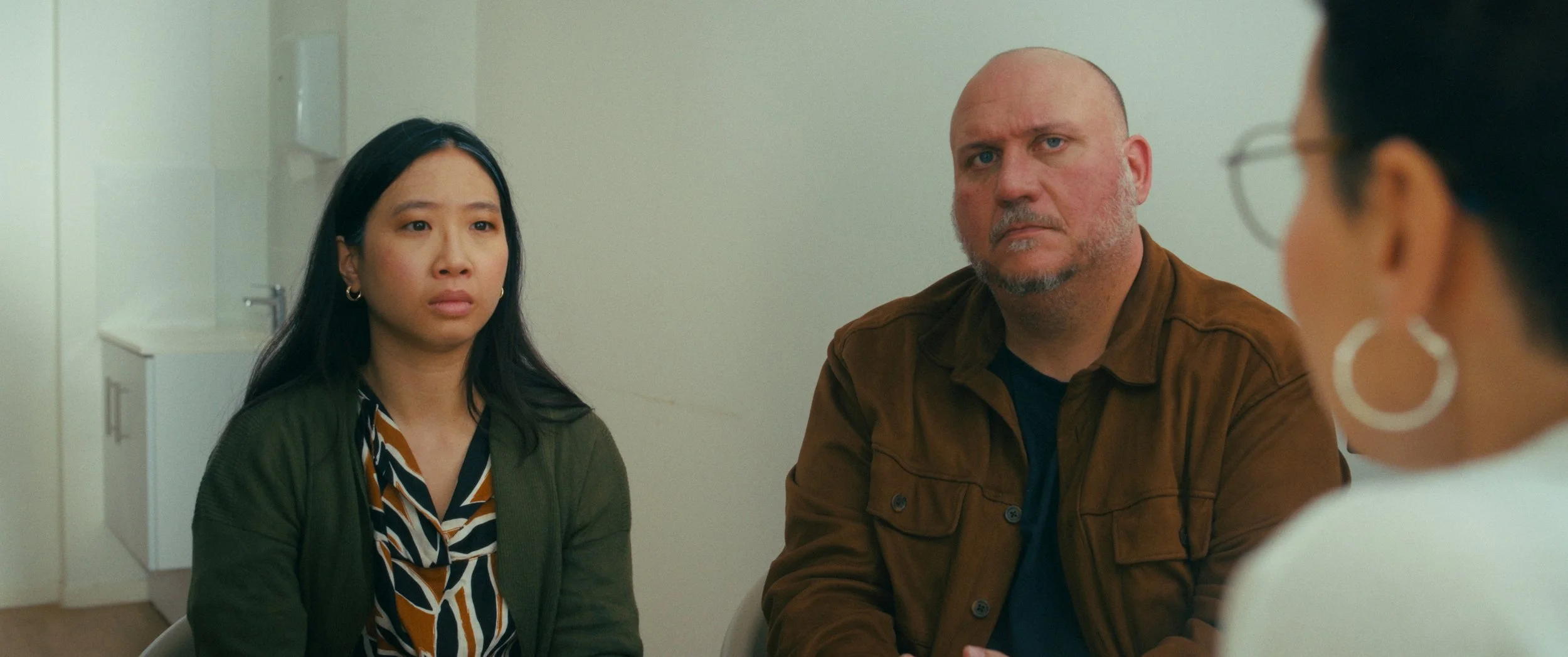 Three people sitting in a room, engaged in a serious conversation. The woman on the left has long black hair, wearing a green jacket and a patterned shirt. The man in the middle has a bald head and a beard, wearing a brown jacket. The woman on the ri