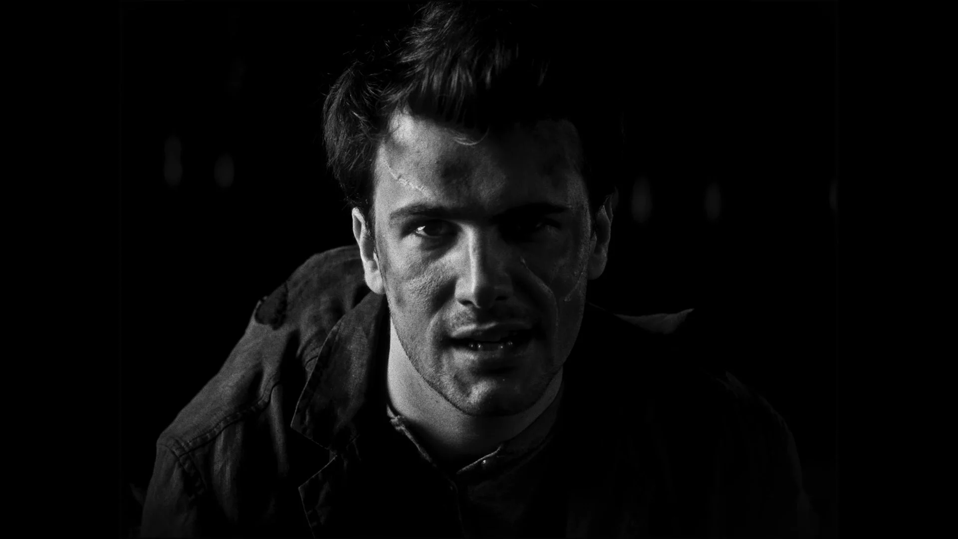 Black and white portrait of a young man with intense expression, slightly messy hair, wearing a jacket, against a dark background.