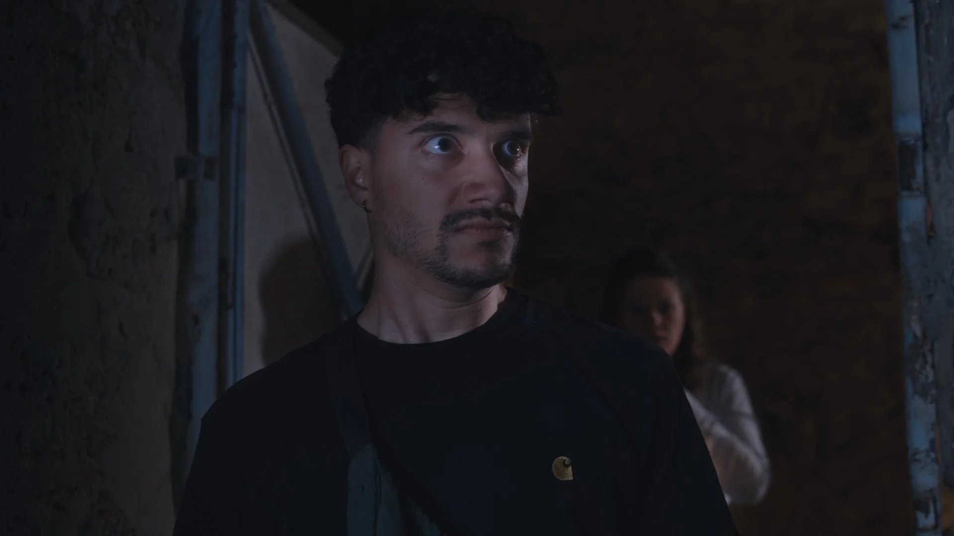 A man with dark curly hair and a beard looks to the left, wearing a black shirt, in a dimly lit setting with a stone wall and a woman in the background.
