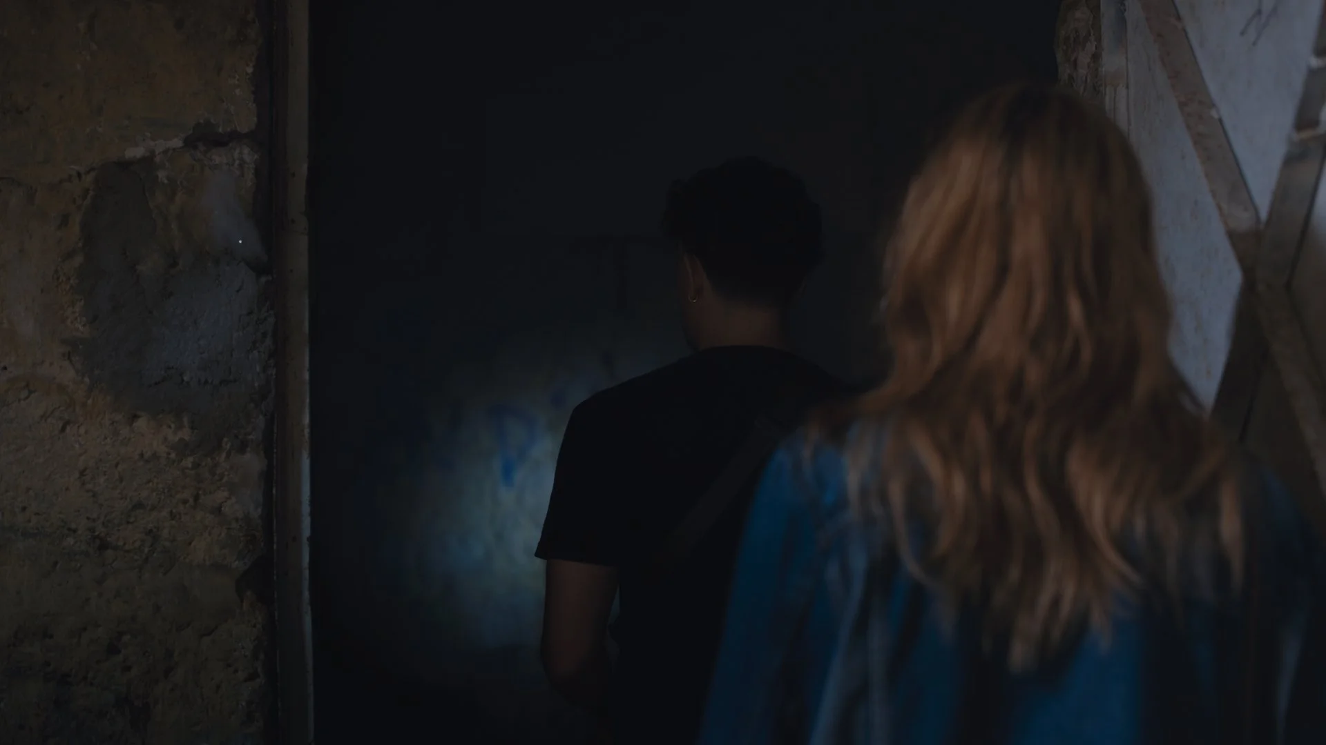 Two people are standing in front of a dark, possibly underground, tunnel or opening. One has long blonde hair and the other has short dark hair. They are seen from behind and facing the opening wall.