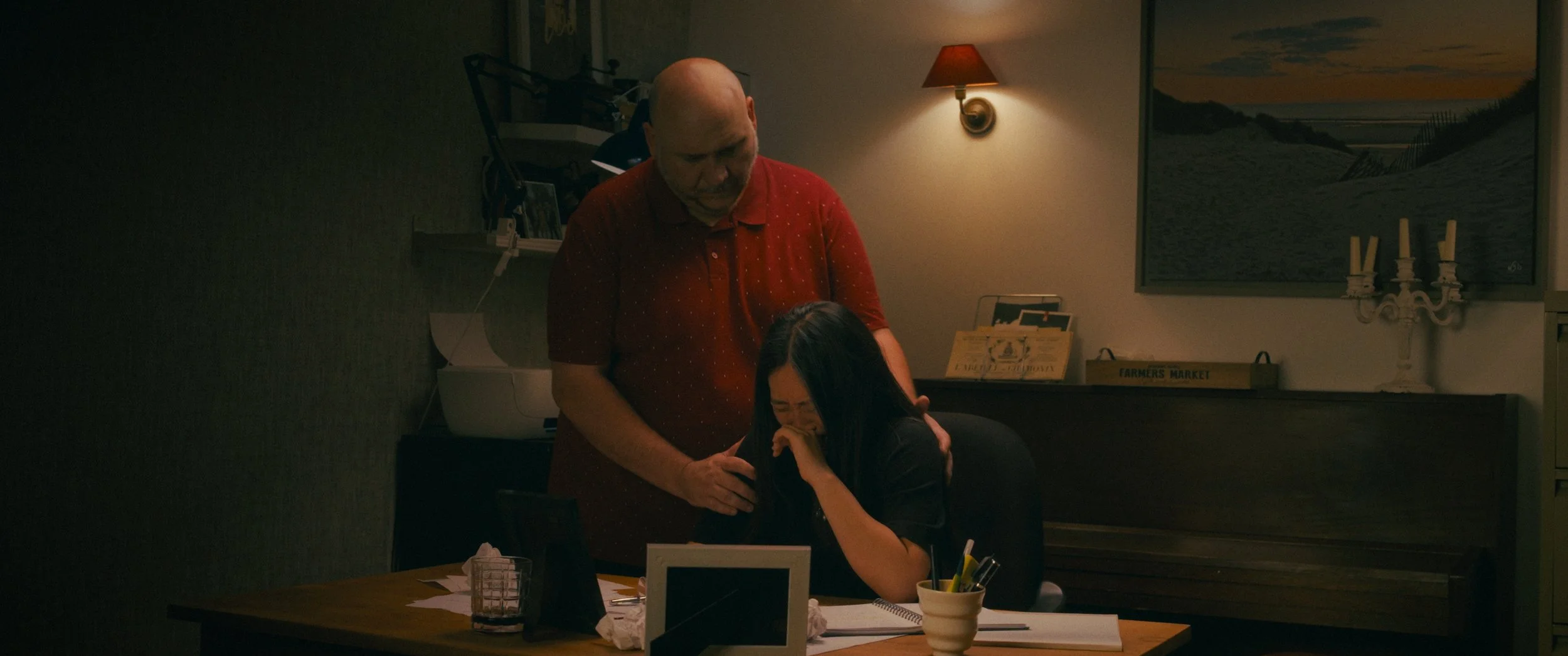 A man standing behind a woman who appears to be upset or crying, in a dimly lit room with a lamp on the wall, a painting of a beach scene, and a table with papers, a cup, and a small picture frame.
